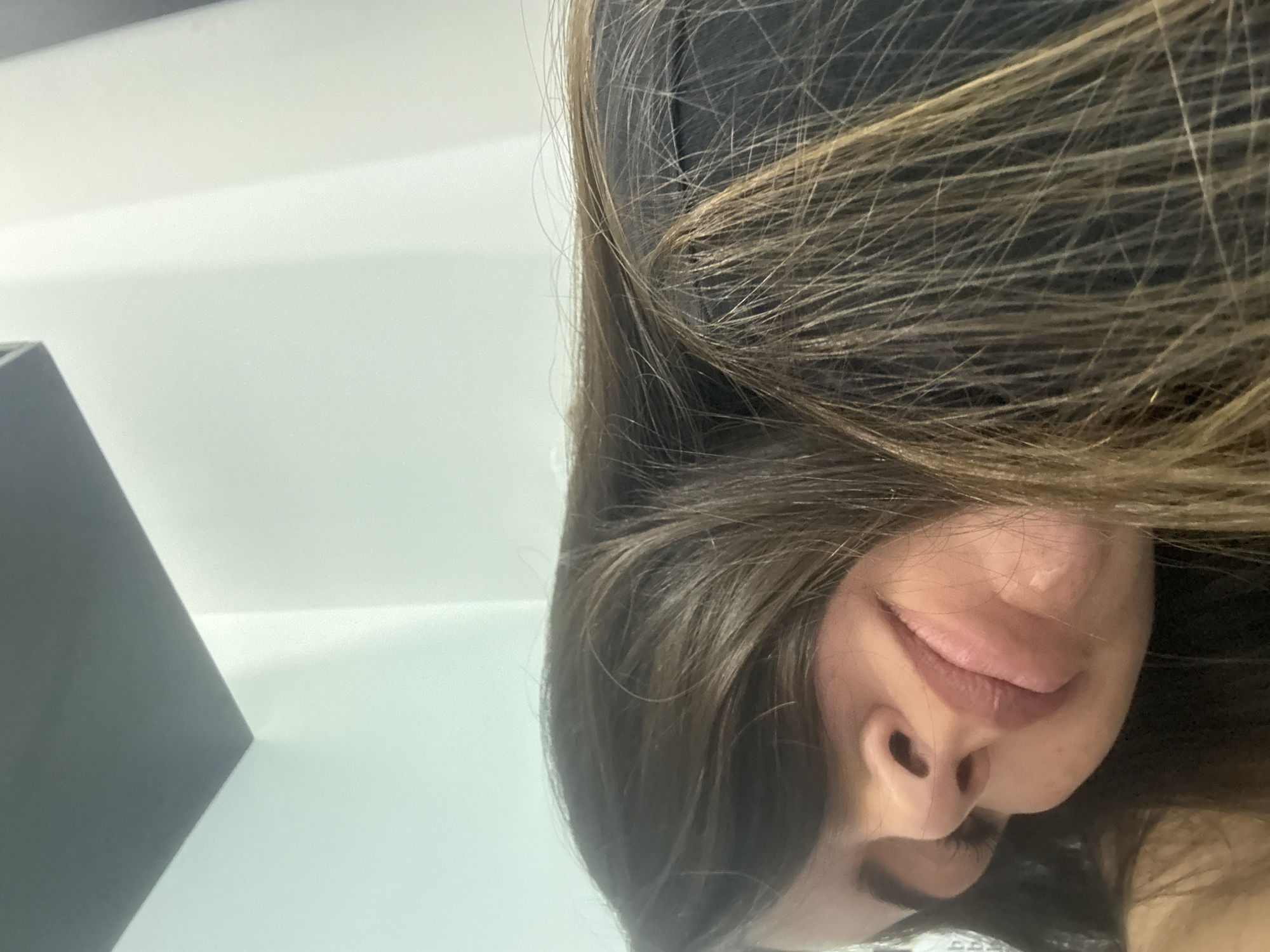 A close-up, tilted perspective shows a woman's face partially obscured by her dark brown hair with blonde highlights. The visible portion of her face includes her nose, mouth with a faint smile, and one closed eye. The background consists of a white wall and a section of a dark gray object, possibly furniture. The location is unknown and not identifiable in the image. There are no indications to suggest it is located in San Bernardino Tlaxcalancingo, Mexico.