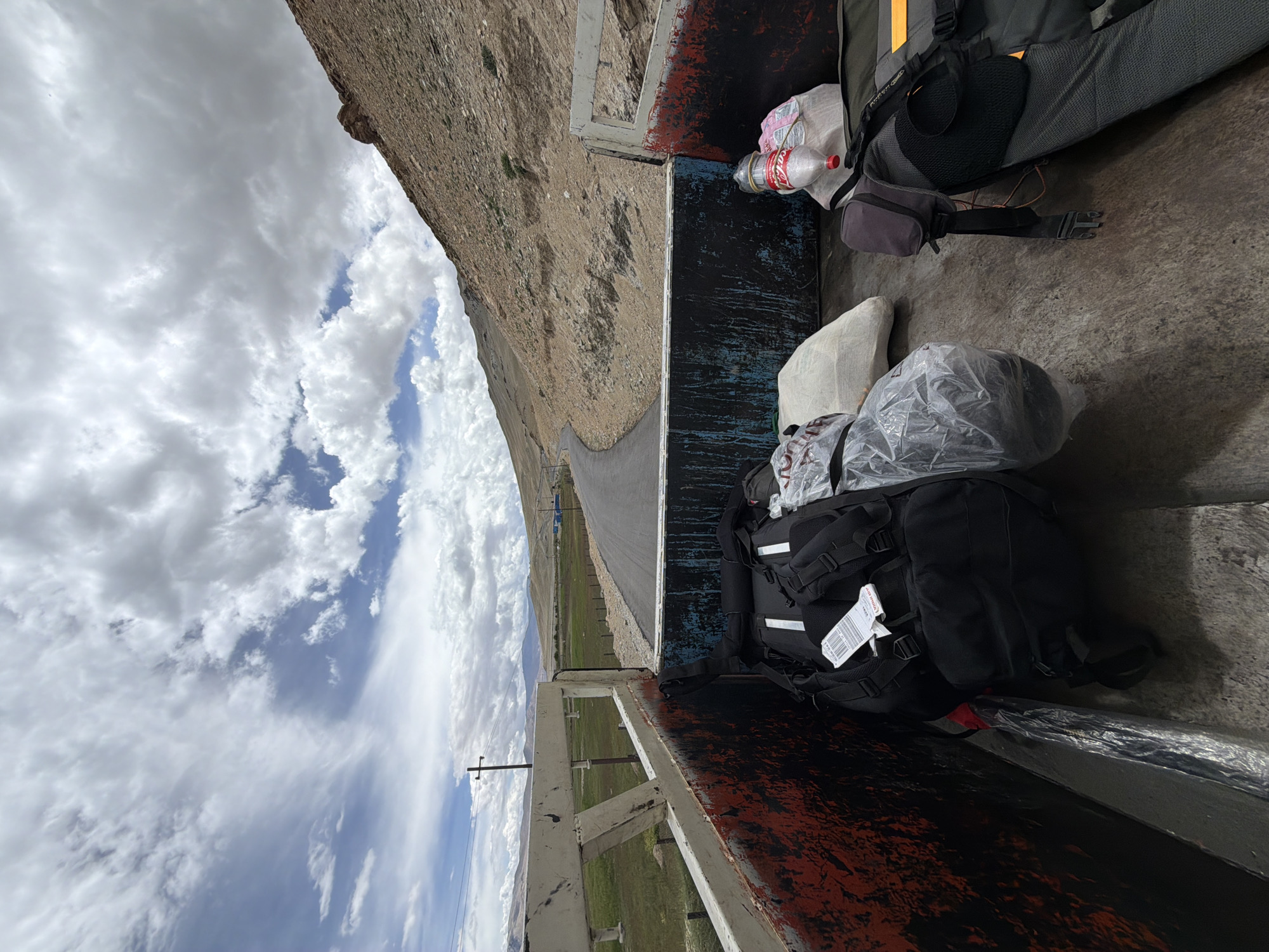 A high-angle, forward-facing outdoor shot shows the back of an open-bed truck filled with luggage. The truck is on a paved road that curves through a dry, mountainous landscape under a cloudy sky. To the left, a grassy field is visible behind a white metal fence and a utility pole. In the truck bed, there are several black backpacks, one clear plastic-wrapped item, a white cloth bag, and a red-labeled water bottle. The truck bed has red and black painted sides. The scene appears to be in Karzok, India, a village located on the shore of Tso Moriri lake in Ladakh.