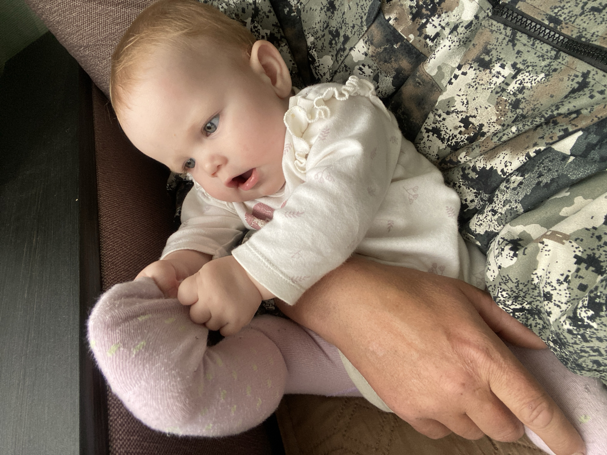 A baby, appearing to be around 6-12 months old, is seated in the lap of an adult. The baby is wearing a white, long-sleeved shirt with ruffled trim, and pink leggings with light green dots. The baby is holding its own foot. The adult is wearing a camouflage patterned jacket or shirt. The setting appears to be indoors, near a dark-colored table and a brown upholstered piece of furniture. There is no verifiable information to suggest this image was taken in the city of Фоки, Russia.