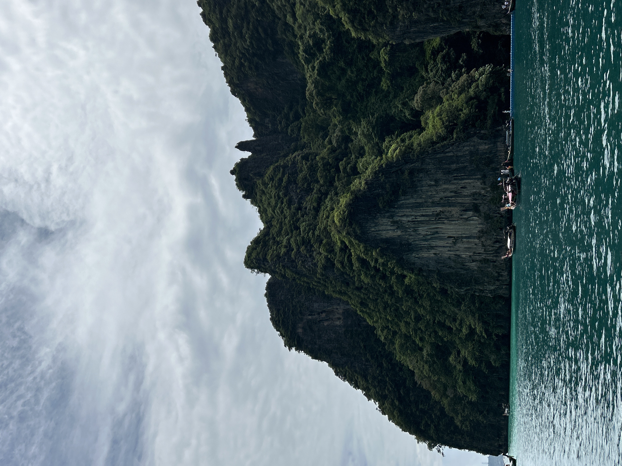 A long-shot photograph captures a body of water with a backdrop of towering, lush green cliffs. The water, a vibrant turquoise, is slightly rippled and dotted with several longtail boats. The cliffs, dense with vegetation, rise dramatically from the water, with some appearing sheer and rocky while others are covered in a thick canopy of trees. The sky above is overcast with a light cloud cover, casting a soft, diffused light on the scene. The overall impression is one of a tropical seascape, likely in a coastal region of Thailand.