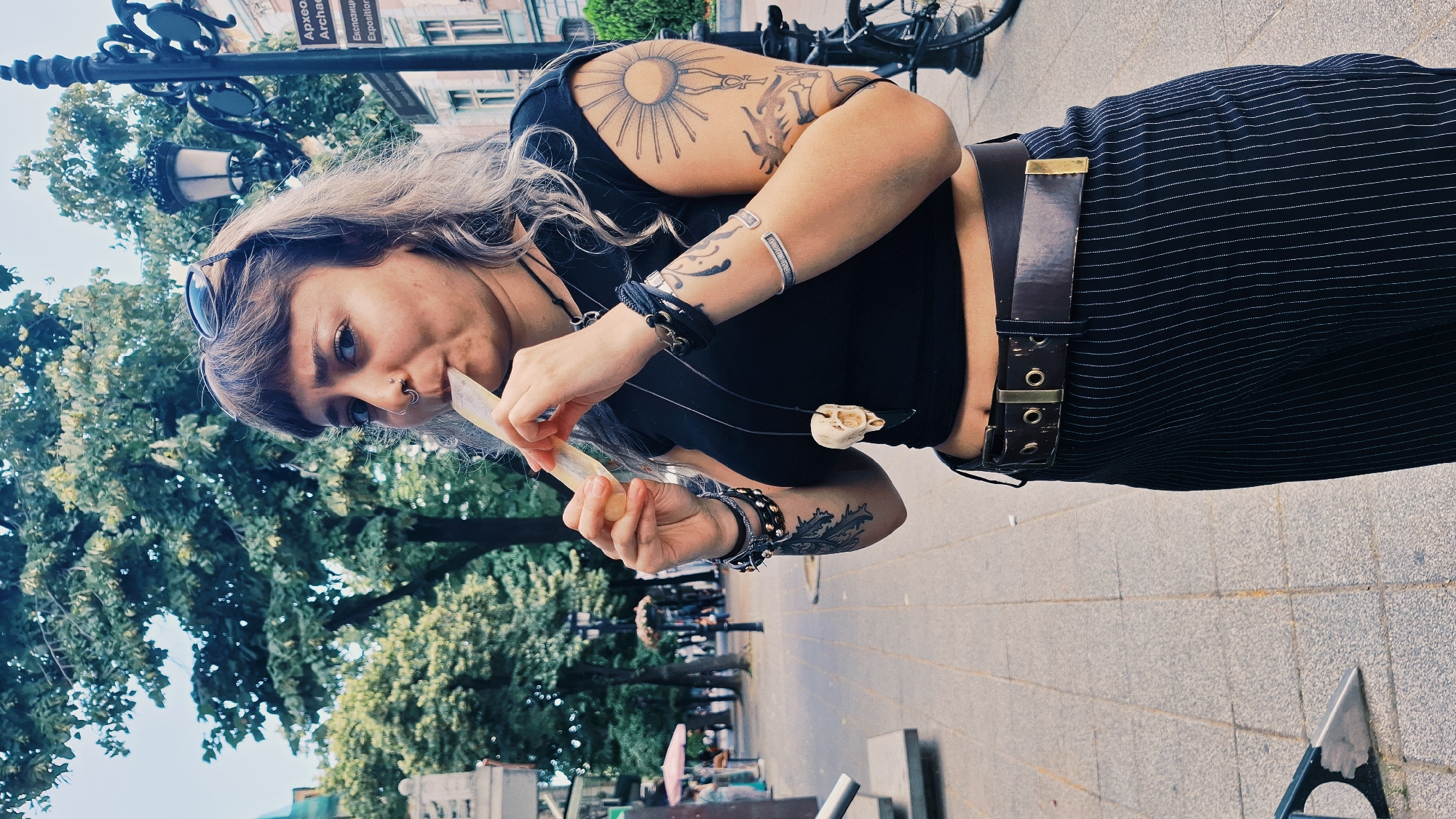 A young woman with long, light-colored hair, tattoos on her arms, and a nose ring is shown in an outdoor, urban setting. She is holding a light-colored object, possibly a piece of paper or currency, to her mouth. She is wearing a black crop top, a dark, pinstriped skirt, and a dark belt with a brass buckle. She also has several bracelets and necklaces, one of which features a small skull charm.
The background shows trees, a street lamp, and buildings, suggesting a city street. The ground is paved with light-colored tiles. The time of day appears to be daytime, with moderate lighting. The overall mood is casual and candid. Some text is visible on a sign in the background, appearing to be in a Cyrillic alphabet, potentially indicating a location in Eastern Europe. One sign reads "Архео" which translates to "Archaeological". The location is identified as Plovdiv, Bulgaria.