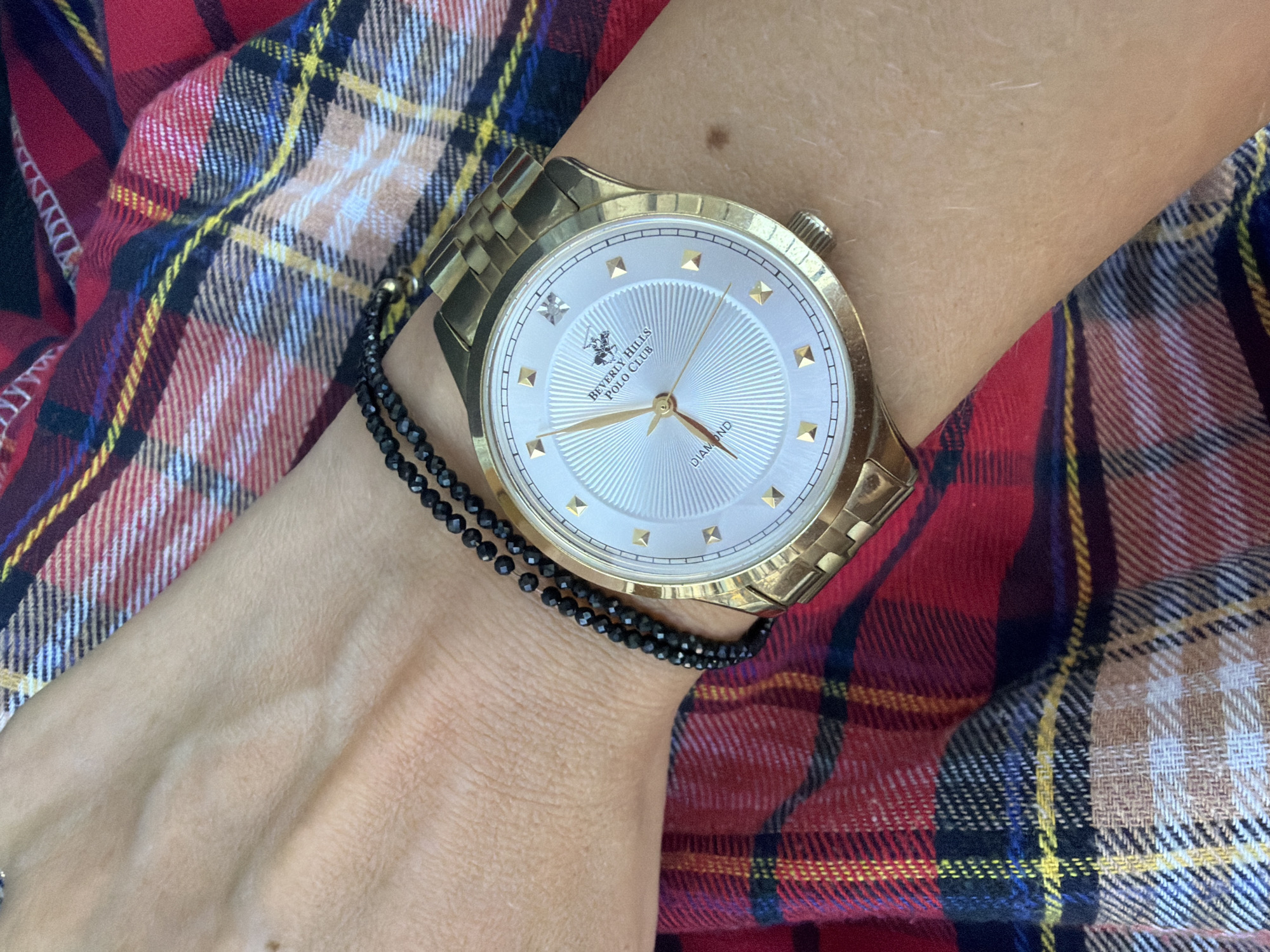 A close-up shot focuses on a person's wrist adorned with a gold-toned watch and a black beaded bracelet, set against a backdrop of red, blue, and yellow plaid fabric. The watch face is white with gold hour markers and a Beverly Hills Polo Club logo. The bracelet is made of small, faceted black beads. The plaid fabric is folded and draped, creating a textured background. The image captures a detail of personal accessories against a patterned textile. The location is Nha Trang Ward, Vietnam.