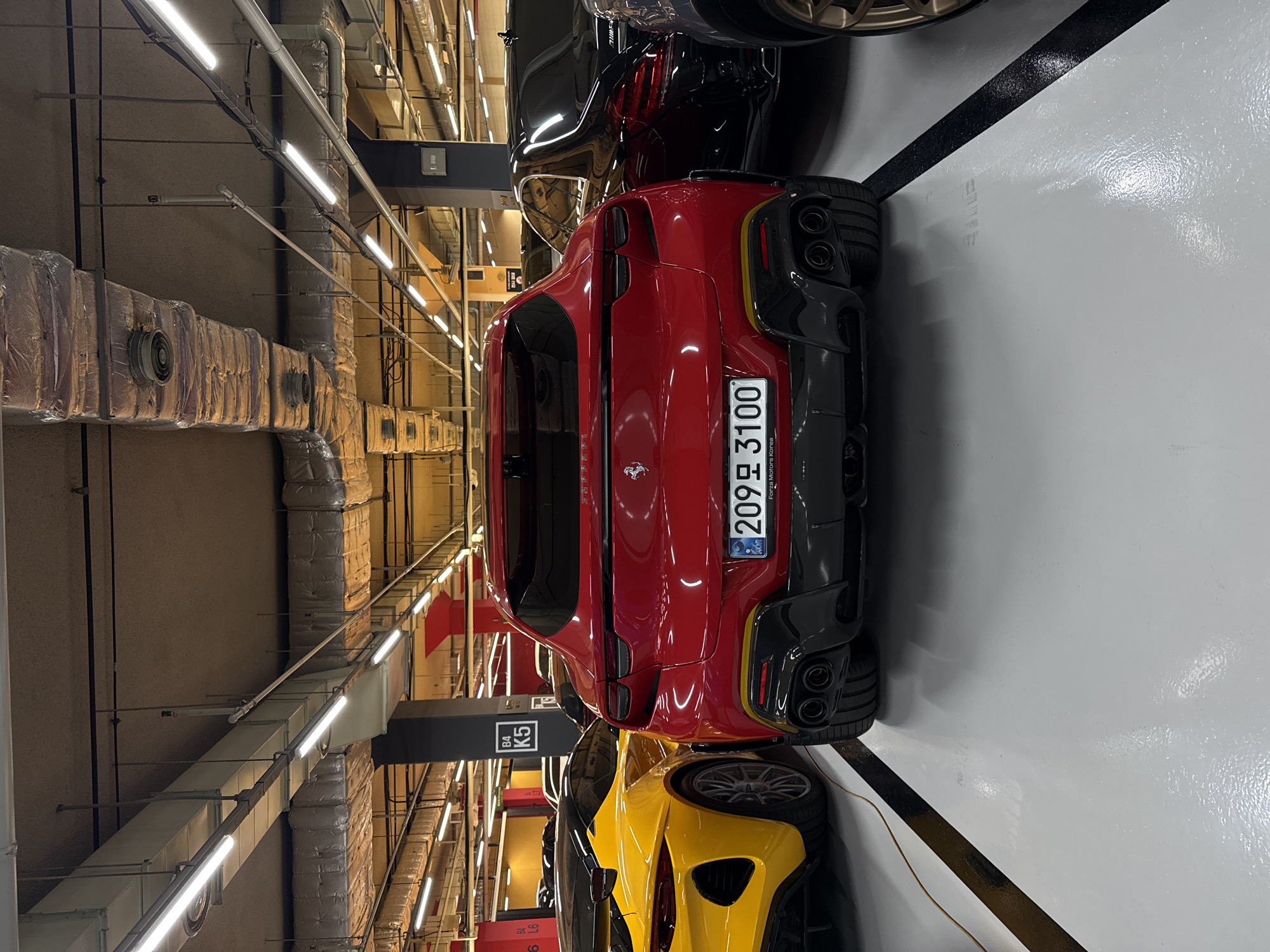 The image shows a low-angle view of a parking garage, likely in Seoul, South Korea, given the license plate visible on the red Ferrari. Several luxury cars are parked closely together, including a red Ferrari, a yellow car, and a black car. The ceiling is equipped with fluorescent lighting and industrial-looking ventilation ducts. The floor is painted white and marked with black lines.