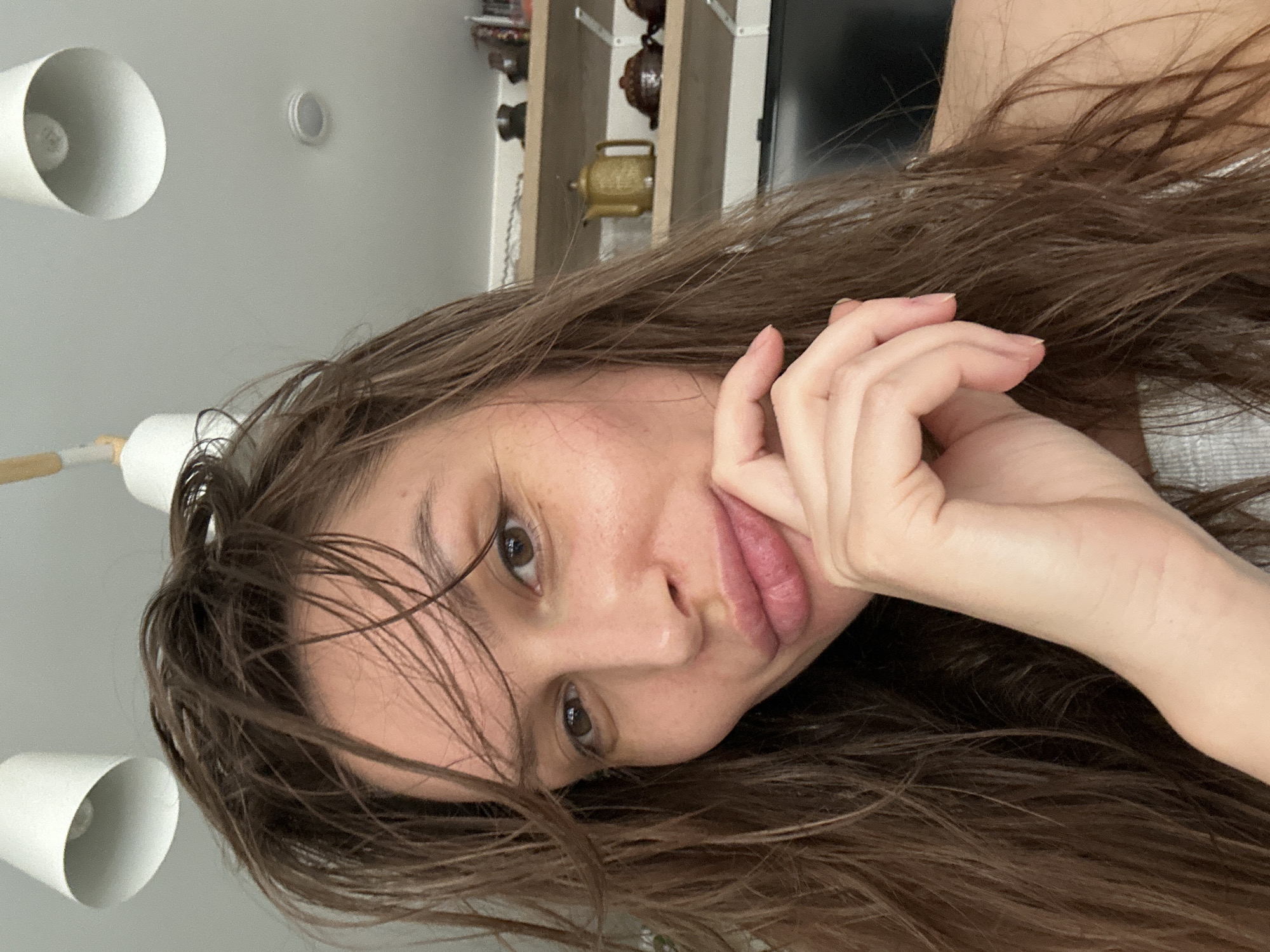 A woman with long, wet brown hair is lying down, looking at the camera. Her right hand is near her face. In the background, a light fixture is visible on the ceiling and a shelf with decorative items is mounted on the wall. The location is Unknown, Unknown.