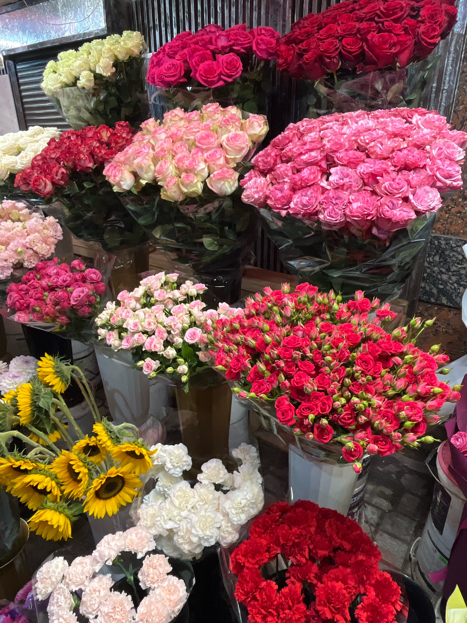 The image shows a close-up, high-angle view of a flower stall in Kyiv, Ukraine, displaying a vibrant assortment of fresh flowers. Dominating the scene are numerous bouquets of roses in various shades, including deep red, bright pink, pale pink, and cream. Interspersed with the roses are bunches of sunflowers with their distinctive yellow petals and dark centers, as well as white and light pink carnations. The flowers are arranged in buckets and wrapped in clear plastic, typical of a market setting. The background suggests an indoor environment with vertical blinds or shelving. The lighting is somewhat dim, highlighting the rich colors of the flowers. There are no people visible in the frame, and the focus is entirely on the floral display.