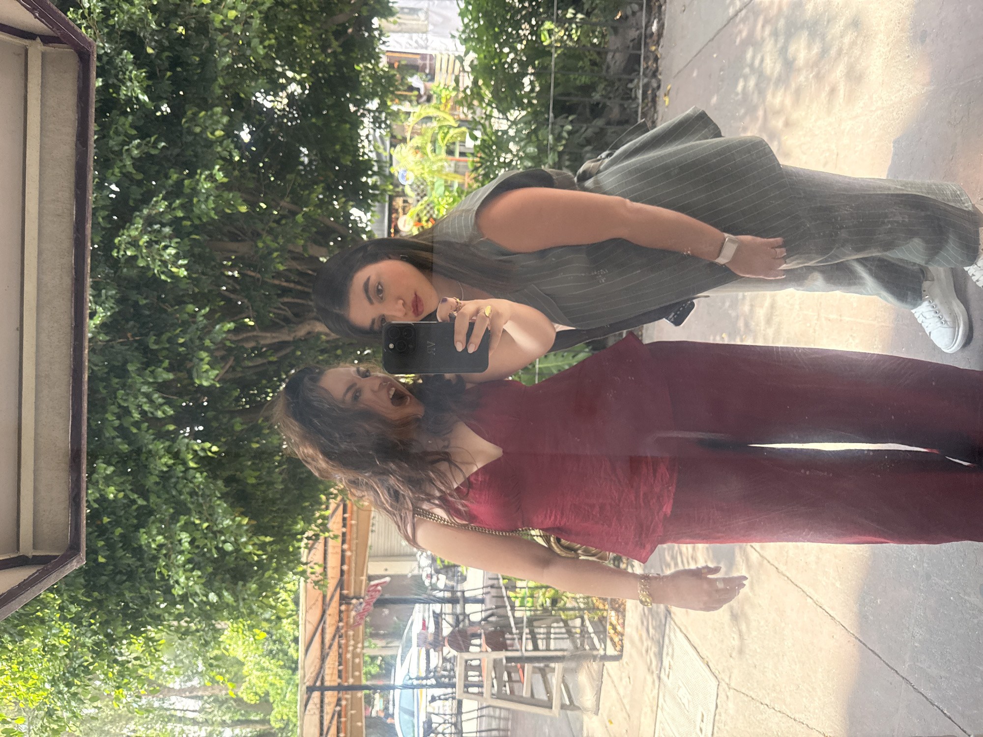 The image depicts two women reflected in a surface, likely a large window or mirror. One woman wears a gray pinstripe suit and white sneakers, holding a smartphone up to take a picture. The other woman, reflected below, wears a red top and matching pants, with gold jewelry on her wrist and neck. Both women are facing forward, towards the camera. Behind them, a lush green background indicates trees and foliage. The reflection also shows a street scene with tables, chairs, and a parked car, suggesting a cafe or restaurant setting. Based on the architectural style and vegetation, the location may be in the city of Mexico City, Mexico. The reflection indicates a sunny day.