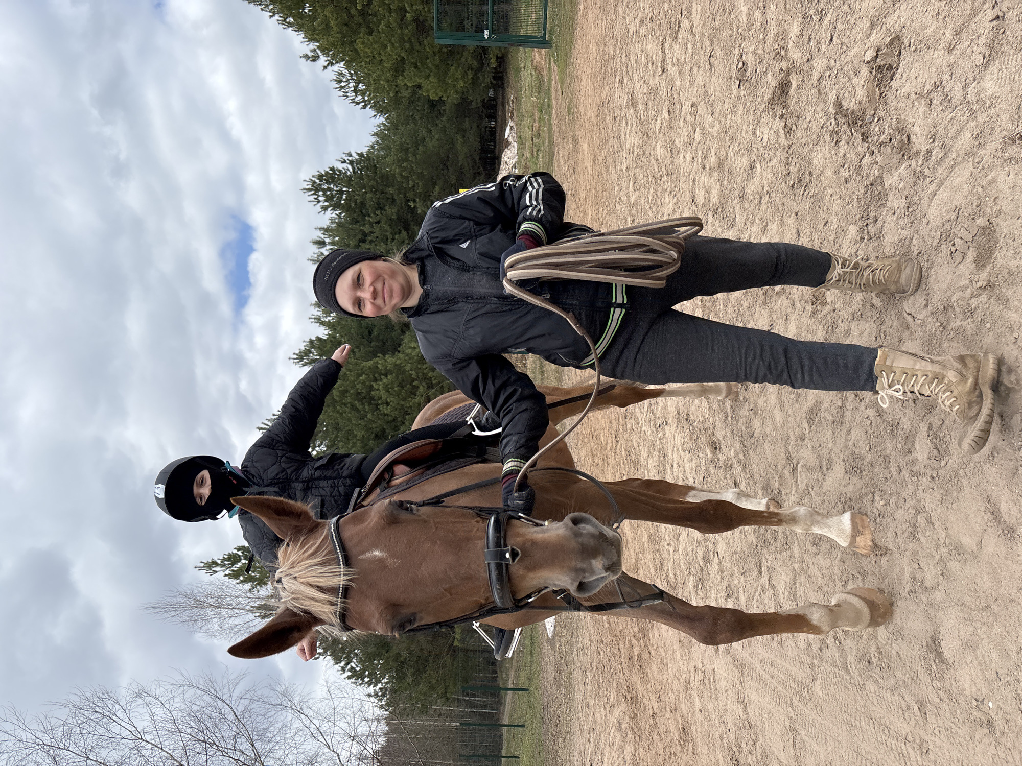The image depicts two individuals and a horse in an outdoor setting in Русский Вожой, Russia. One person, wearing a black jacket, grey pants, and tan boots, stands next to a brown horse and holds coiled reins. This individual wears a black beanie with possible writing on the front, and their clothing features Adidas-style stripes. A second person, wearing a black ski mask and a black jacket, is seated on the horse, raising one arm. The horse has a light blonde mane. The background includes bare trees and a dense pine forest under a cloudy sky. The ground is sandy. A metal gate is visible in the distance.
