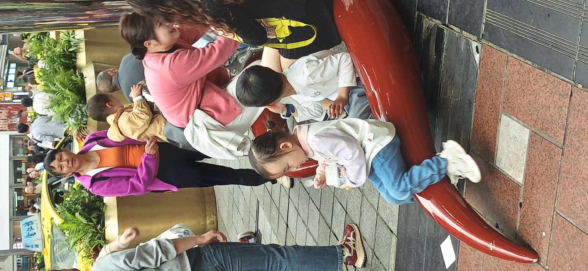 The image captures a street scene in Yuzhong District, China. Several people, including adults and children, are present. A woman in a pink sweatshirt sits next to a person in a black shirt with a yellow design. Near them, a young boy in a white shirt looks down. Adjacent to these individuals, a little girl in a light-colored sweater and jeans sits on a red, curved object, possibly a sculpture or part of street furniture, and appears to be eating something from a container. Another woman in a purple jacket stands nearby. The scene is set on a tiled and paved pedestrian area, with buildings, greenery, and a yellow taxi visible in the background, suggesting a bustling urban environment. A crowd of people can be seen in the distance. The objects include containers, a spoon, and shoes. The actions include sitting, eating, and interacting.