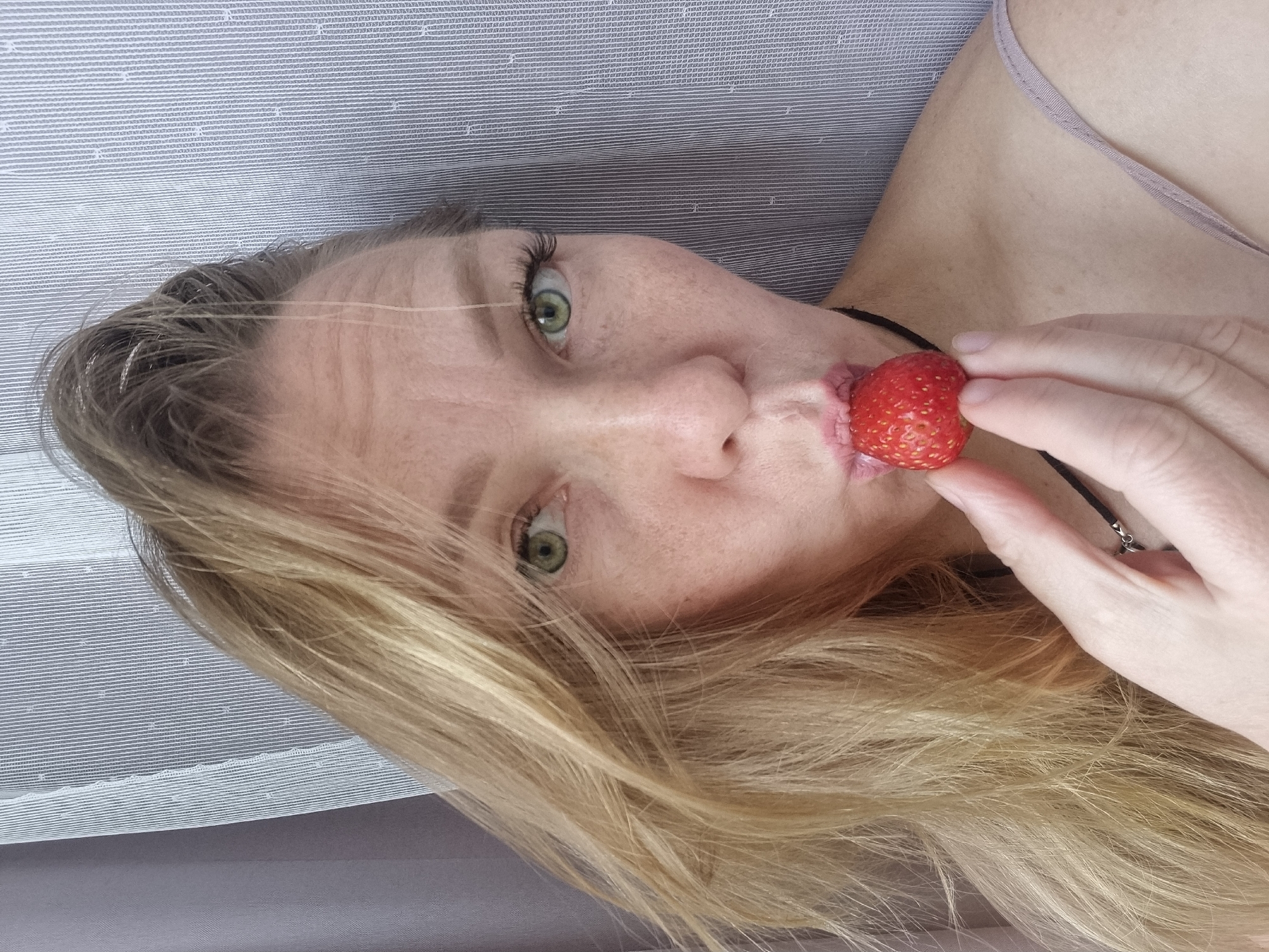 A woman with blonde hair and green eyes is holding a strawberry to her lips. She is wearing a purple top and a black necklace. The background is a patterned curtain. The location is Chaikovsky, Russia.