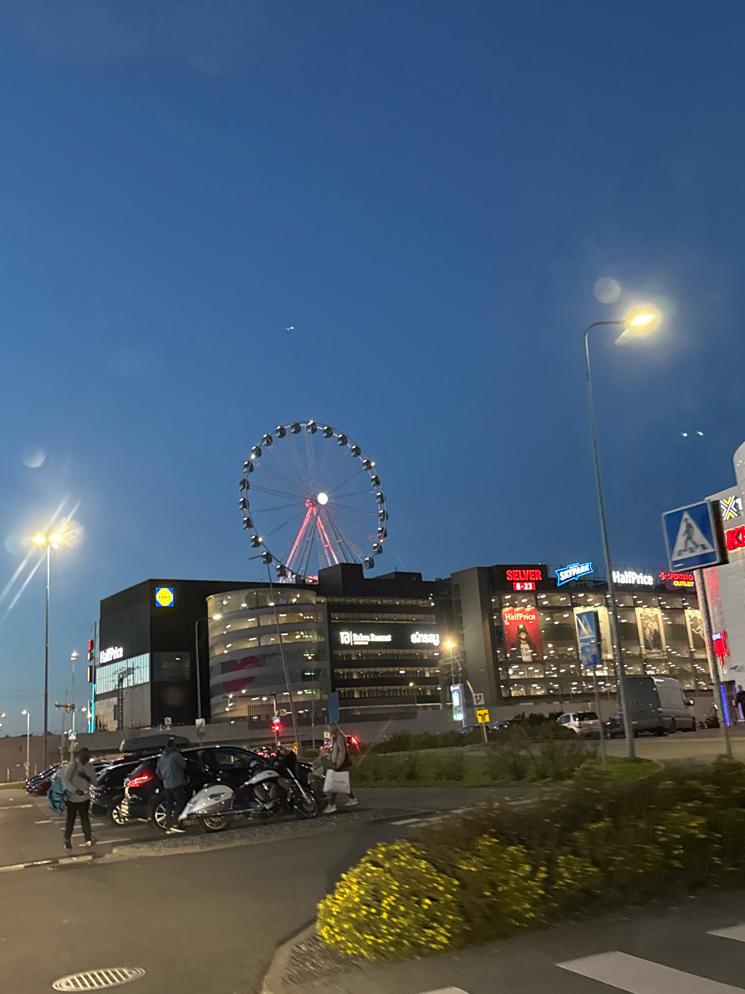 The image shows a modern urban scene at dusk in Tallinn, Estonia. A large Ferris wheel, illuminated with red and white lights, dominates the background, suggesting a recreational area or a city landmark. Adjacent to the Ferris wheel are several multi-story buildings with illuminated windows and prominent signage, including "Lidl," "HalfPrice," and "Selver," indicating a commercial district, likely a shopping center or retail park. The sky is a deep blue, transitioning from twilight to night. Streetlights are visible, casting a warm glow. In the foreground, several cars and a motorcycle are parked on a paved area, possibly a parking lot or a street. A few individuals are present, some interacting with the vehicles. The overall environment appears to be well-lit and functional, characteristic of a developed urban area.