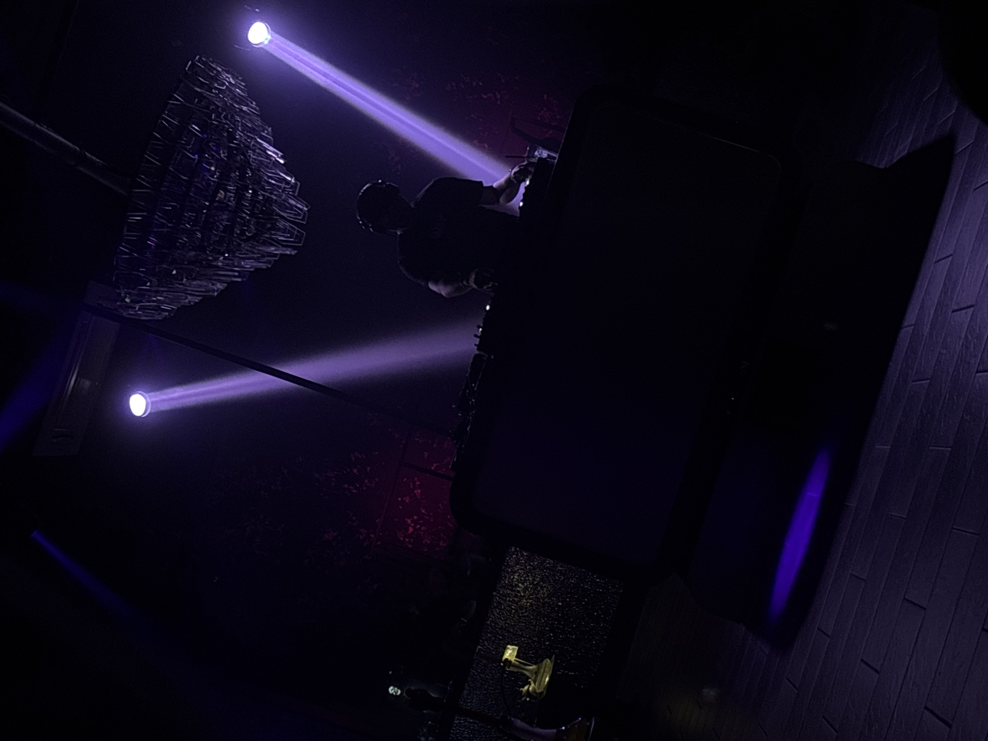 This image depicts a dimly lit, indoor scene, likely a concert venue or nightclub. The primary subject is a person, silhouetted against purple lighting, wearing headphones and a dark t-shirt. They appear to be operating DJ equipment or a similar console, indicated by their posture and outstretched hands. The overall atmosphere is moody and energetic, suggested by the dramatic purple spotlights and the dark, textured walls and floor.
In the background, a large, angular, dark object, possibly a stage setup or a piece of furniture, dominates the right side of the frame. Above and to the left, an intricate, geometric light fixture hangs, catching the purple light. Two bright purple beams of light cut through the darkness from above, creating a theatrical effect. The floor is tiled with dark wood planks, and a small, golden object, possibly a trophy or a decorative item, rests on the floor in the lower center. The scene is devoid of any visible text, and the time of day is indeterminate, but the lighting suggests nighttime or a dark indoor environment.