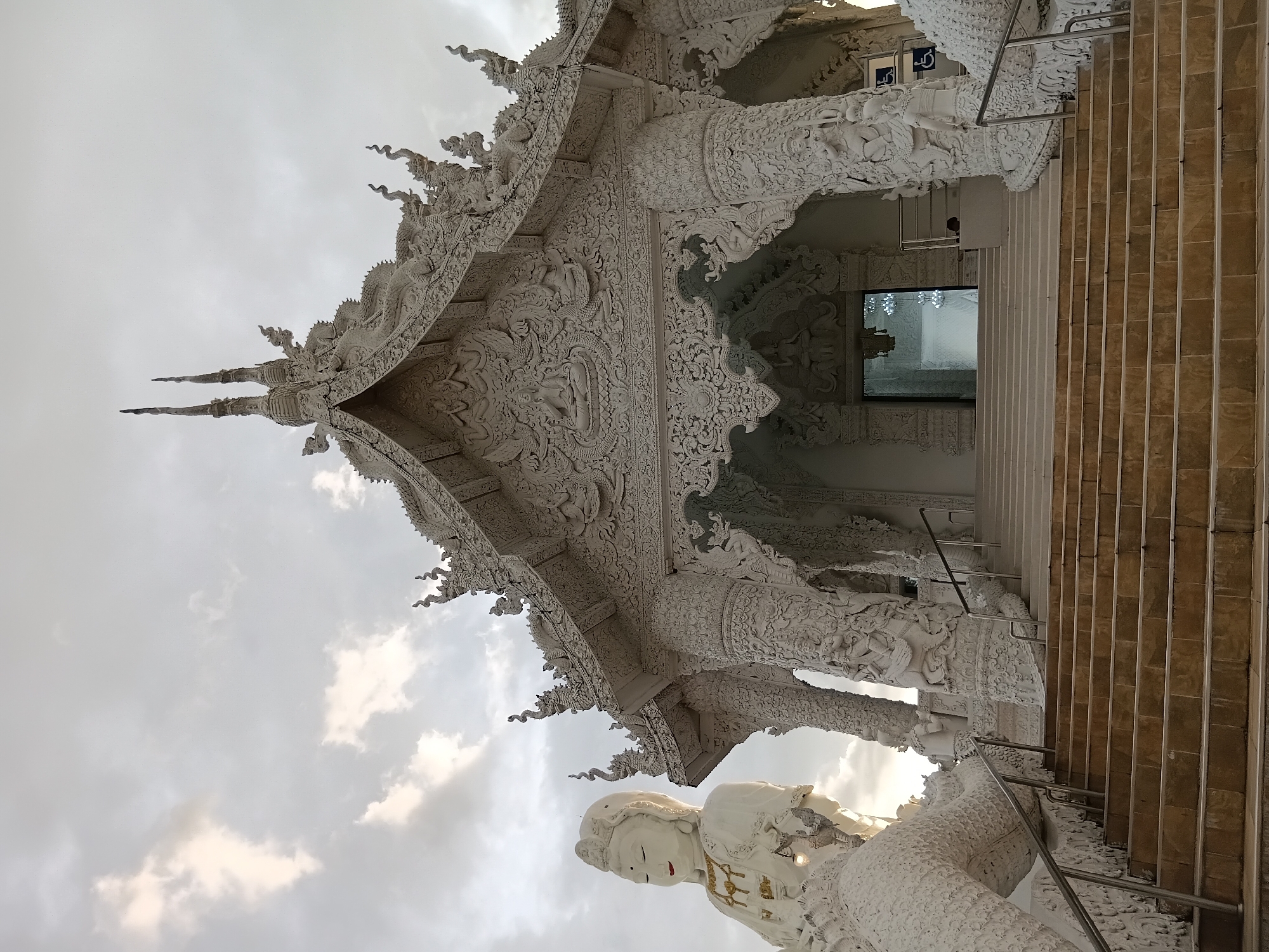 The image shows the exterior of a white Buddhist temple, likely Wat Rong Khun (The White Temple) in Chiang Rai, Thailand, which is near Ban Lao Phatthana. The temple is elaborately decorated with intricate carvings and sculptures. In the foreground, a wide set of stairs leads up to the temple entrance. To the left of the stairs, a large white statue of a seated figure, possibly Guanyin, is visible. The sky above is overcast with dramatic clouds. The overall impression is one of awe-inspiring and detailed religious architecture.