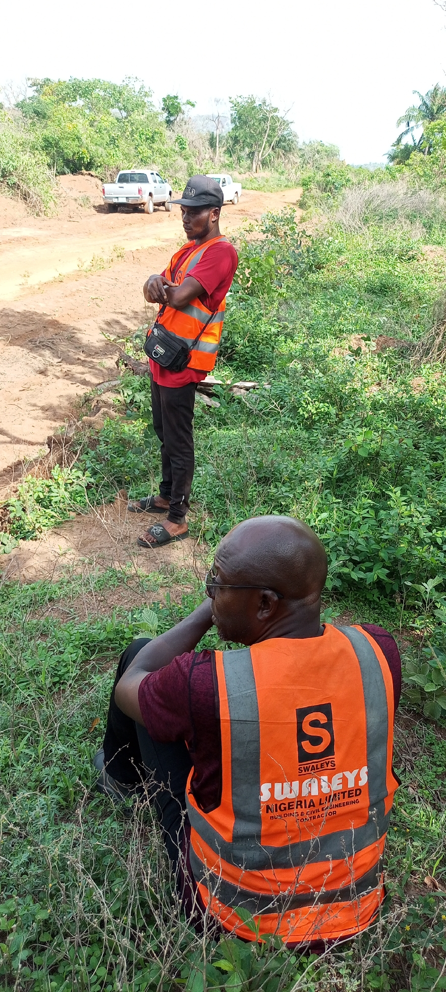 Two men are depicted outdoors in what appears to be a rural or undeveloped area in Otunade, Nigeria. The scene features lush green vegetation, dirt tracks, and scattered trees under a bright, hazy sky.
In the foreground, a bald man with glasses is sitting or crouching, viewed from behind and the side. He wears a maroon t-shirt and an orange reflective vest. Visible on the back of his vest is a black logo featuring a large 'S', with the text "SWALEYS" above "SWALEYS NIGERIA LIMITED BUILDING & CIVIL ENGINEERING CONTRACTOR". He is looking towards the right side of the frame, seemingly resting or observing.
Behind him, another man stands upright, his arms crossed. He wears a red t-shirt, an identical orange reflective vest, black pants, and dark sandals. A black cross-body bag hangs across his chest. He also wears a black cap with the letters "OLO" visible on it. His gaze is directed to his right, giving an impression of observation or contemplation.
In the background, on a dirt road, two pickup trucks are parked or driving slowly. One appears silver/grey, and the other is white. The presence of the reflective vests and the company name on them suggests that the men are likely involved in surveying, construction, or civil engineering work in this undeveloped locale. The abundant greenery and dirt pathways indicate an environment that is not highly urbanized. The time of day appears to be bright daylight.