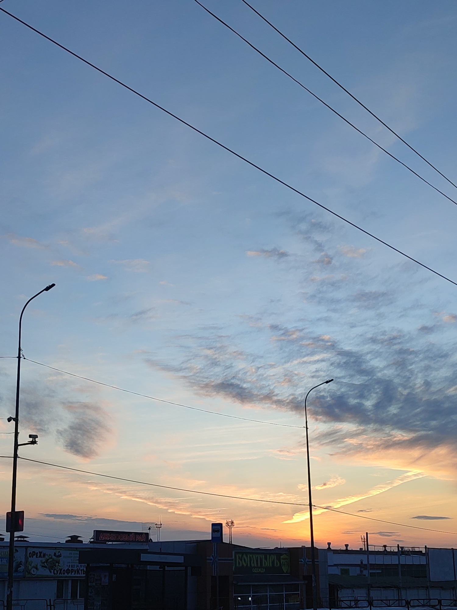 The image captures a sunset in the city of Chaikovsky, Russia. The sky is a mix of blues, pinks, and oranges, with streaky clouds. Several power lines cross the upper part of the frame. Two streetlights stand on either side of the image.
In the lower part, buildings with storefronts are visible. On the left, there is a shop sign that reads "ОРЕХИ СУХОФРУКТЫ," translating to "Nuts Dried Fruits." Further to the right, the sign "ДЖИНСОВЫЙ РАЙ" (translated to "Jeans Paradise") is noticeable. On the right, a shop is labeled "БОГАТЫРЬ ОДЕЖДА" which means "Hero Clothes". In front of the buildings, there is a railing and snowy ground.