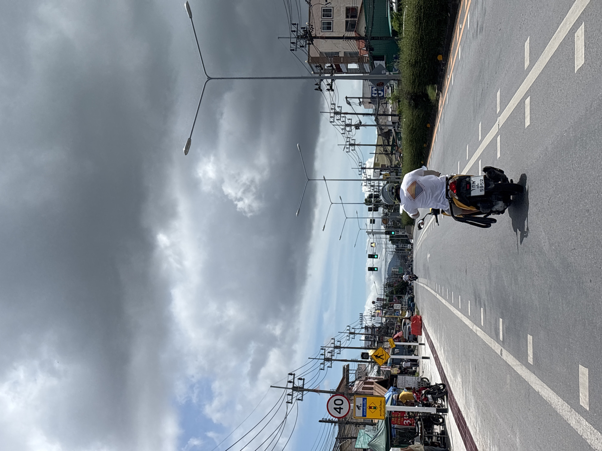 The image depicts a street scene in Rawai, Thailand, under a cloudy sky with patches of blue. In the foreground, a person is riding a yellow scooter from the perspective of someone behind them, wearing a white t-shirt and a helmet. The scooter has a license plate with Thai script and numbers. The road is asphalt with white lane markings. Along the sides of the road are buildings, power lines supported by poles, streetlights, traffic signals, and various signs. On the left side of the road, there are market stalls with awnings and some people standing near them. One sign indicates a speed limit of 40. The overall impression is a busy urban street with typical infrastructure and activity.