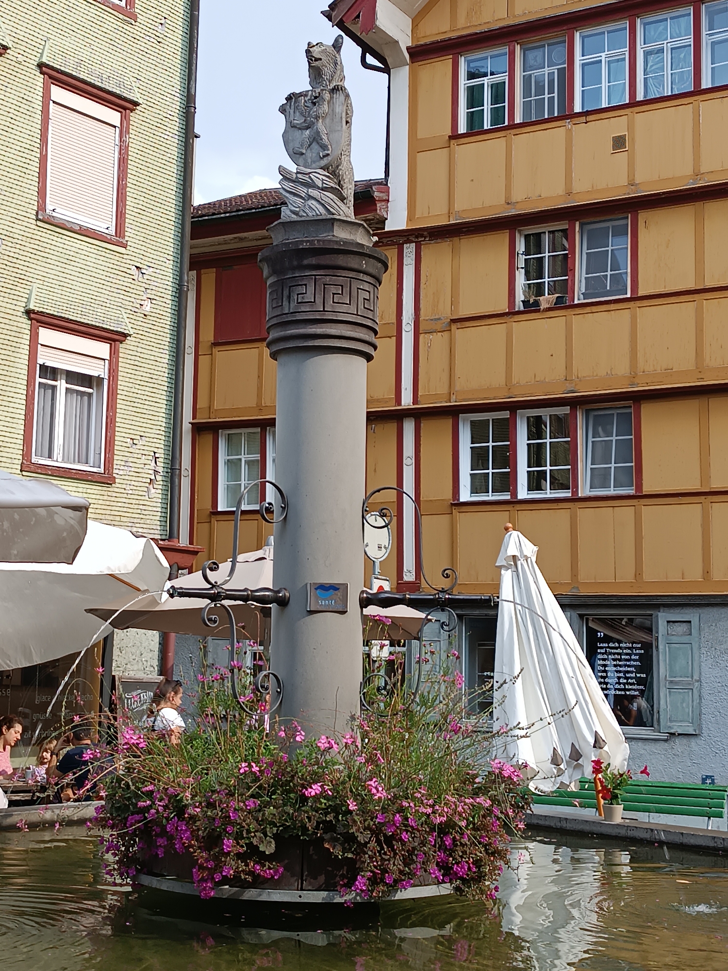 This is a medium shot, taken from a low angle, of a public fountain in the town square of Appenzell, Switzerland. The fountain features a stone pillar topped with a bear statue holding a shield, a common symbol for the region. The pillar is adorned with a Greek key pattern. The base of the fountain is filled with vibrant pink and purple flowers and sits in a pool of water. In the background, traditional Appenzell buildings with colorful facades and distinctive window frames are visible. The building to the left has a pale yellow tiled exterior, while the building to the right is painted ochre with red trim. Several people are seated at tables and on a bench near the fountain, some appearing to be patrons of a nearby cafe. A large white umbrella is propped up near the buildings. The overall scene depicts a lively outdoor space in a Swiss town, suggesting a sunny day.
