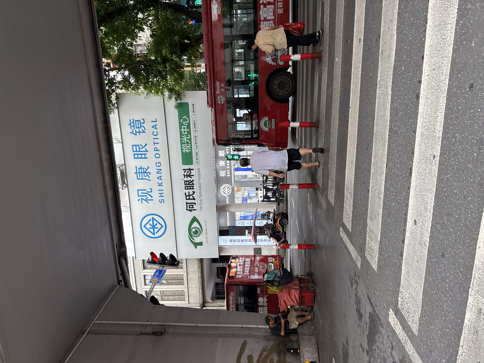 This image depicts a street scene in Yuexiu District, China, under an elevated roadway or structure. A red bus is prominent in the middle right of the frame, with its passengers visible through the windows. Several individuals are present: a man and woman walking across a pedestrian crossing in the foreground, a man seated on the curb on the left with a red cart and belongings, and another person on a motorized tricycle under an umbrella. Overhead, signage for "SHI KANG OPTICAL" and "He Eye Specialist Hospital" is visible. The street is paved, with distinct white pedestrian crossing stripes. Traffic lights and street lamps are also present. Trees are visible in the background.
