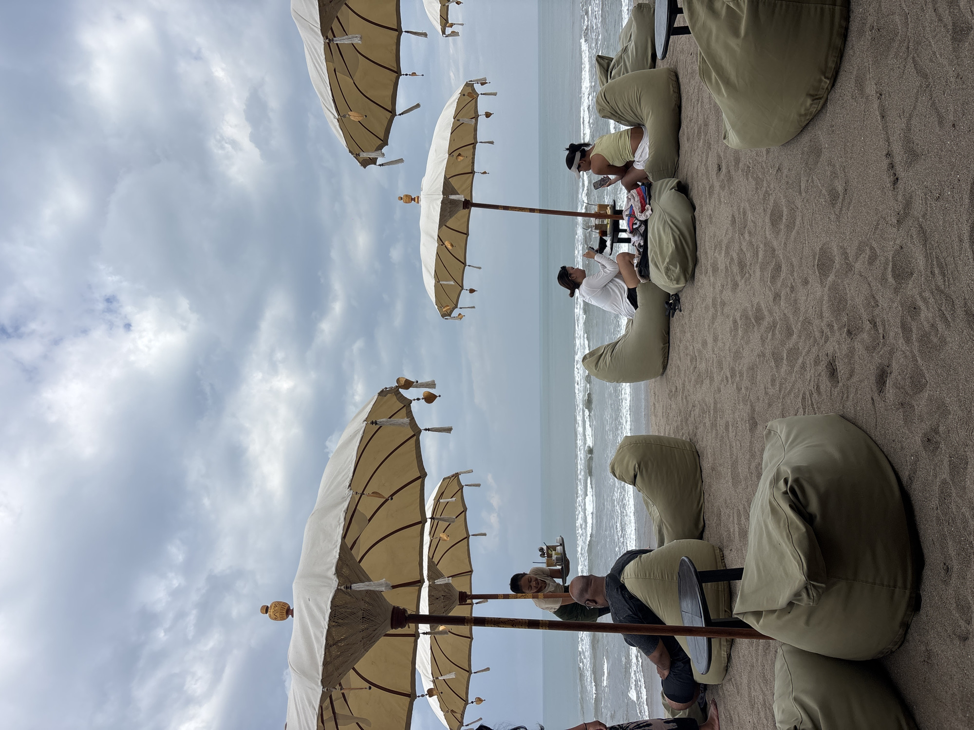 A beachside seating area in North Kuta, Indonesia, features numerous large, olive-green beanbag chairs arranged on dark sand. Several large, cream-colored umbrellas with decorative tassels provide shade. In the background, the ocean stretches to the horizon under a cloudy sky, with moderate waves breaking on the shore.
Several people are present:
- A server, appearing to be a young woman, is carrying a tray with drinks towards two individuals seated at a low table. She is smiling.
- Two individuals are seated at a low table, one on a beanbag and another on a chair. They are looking at their phones.
- A man is seated on a beanbag chair, facing away from the camera, with his back to the ocean.
- Another person is partially visible on the far left, also on the sand.
The scene suggests a relaxed, resort-like atmosphere. The beanbag chairs are scattered around small, dark, circular tables. The umbrellas are prominent features, adding to the ambiance. The overall lighting is diffused due to the overcast sky.