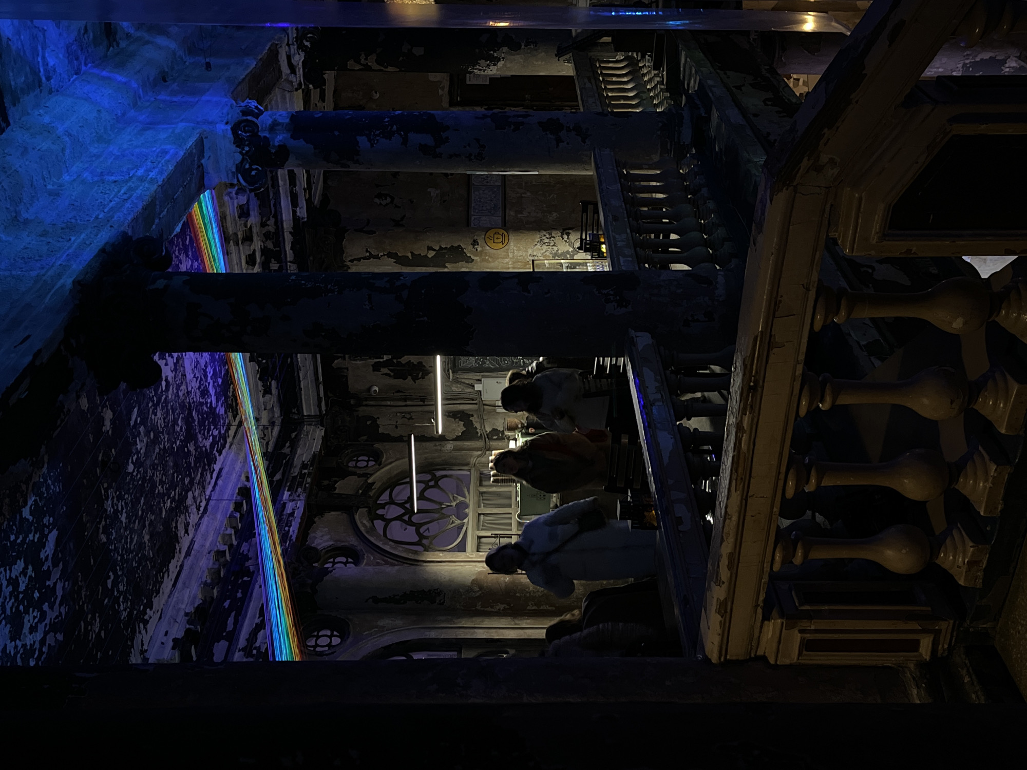 The image depicts the interior of a large, multi-level building at Sevcable Port on Vasilyevsky Island in Saint Petersburg, Russia. The scene is characterized by dim, artificial lighting and a distressed architectural aesthetic.
The environment features multiple classical columns with fluted shafts and ornate capitals, exhibiting extensive paint peeling and discolored surfaces. High ceilings with visible decorative moldings and walls throughout the space show significant paint degradation and exposed layers. Large, arched windows with intricate circular patterned grilles are visible in the background.
Lighting includes a prominent horizontal LED strip displaying a rainbow spectrum, running across the upper section of the room near the ceiling. A substantial portion of the ceiling and an upper wall area are illuminated by a distinct blue light. Functional fluorescent tube lights are visible in the background. A yellow circular sign depicting a coffee cup with steam is mounted on a distant wall, highlighted by a focused light source.
In the mid-ground, approximately five to six individuals are present. Three women are central: one in a light-colored coat carrying a shoulder bag, one in a warm-toned jacket, and another in a light top. They are standing, appearing to observe the surroundings or converse. Their attire is casual. Tables and chairs are partially visible, suggesting a cafe or social area.
The foreground features a weathered balustrade with multiple balusters, showing chipping paint and wear, suggesting a balcony or an upper landing. The perspective is from this elevated position, looking across the space. No overt actions or specific interactions are discernible beyond general presence within the environment.