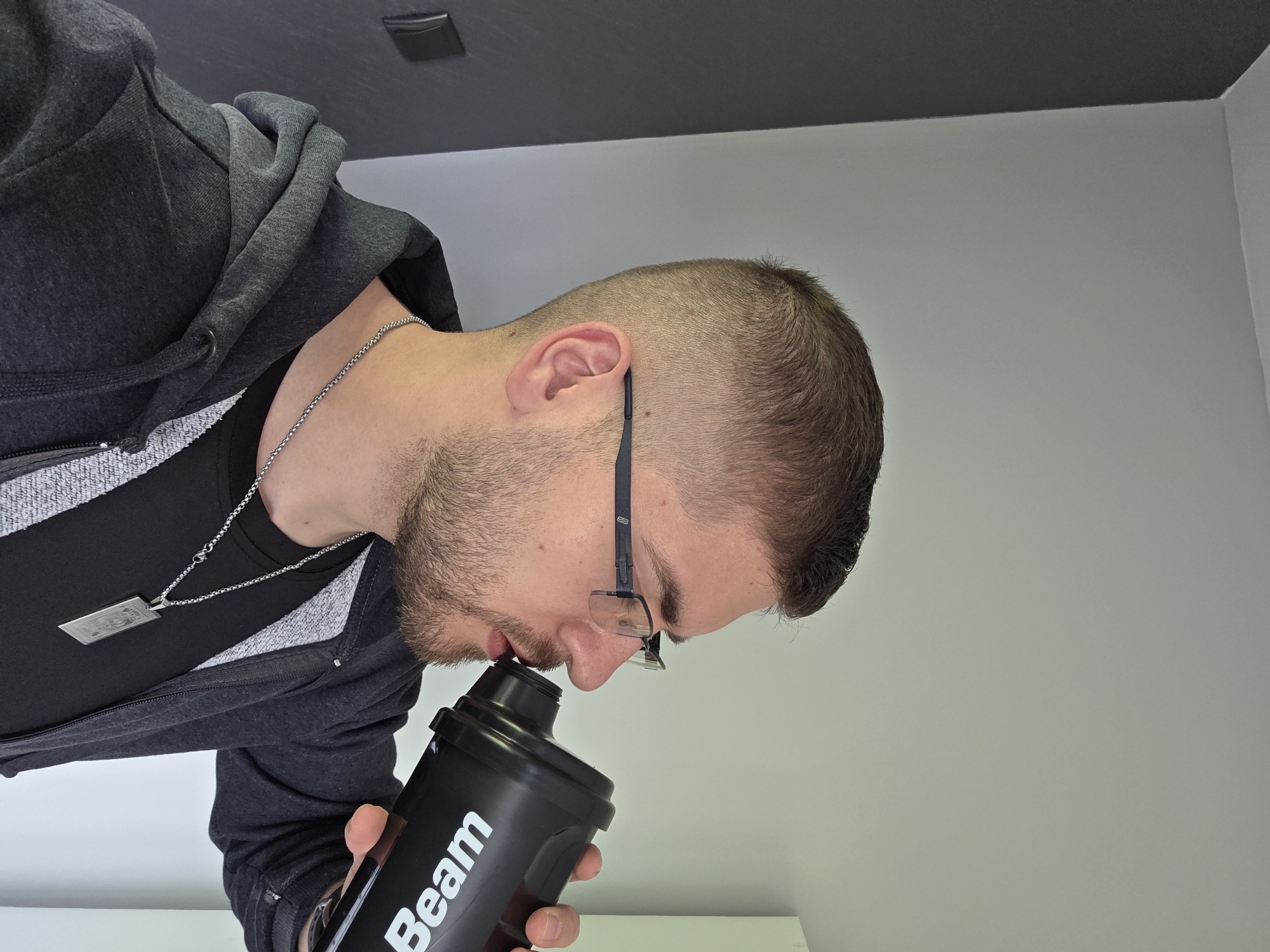 A man is drinking from a black "Beam" shaker bottle. He has short brown hair, a beard, and is wearing glasses, a black t-shirt, and a gray hoodie with a silver necklace. The background consists of a black ceiling, a white wall, and a white shelf. The location cannot be determined to be Yagodovo, Bulgaria from the given image.
