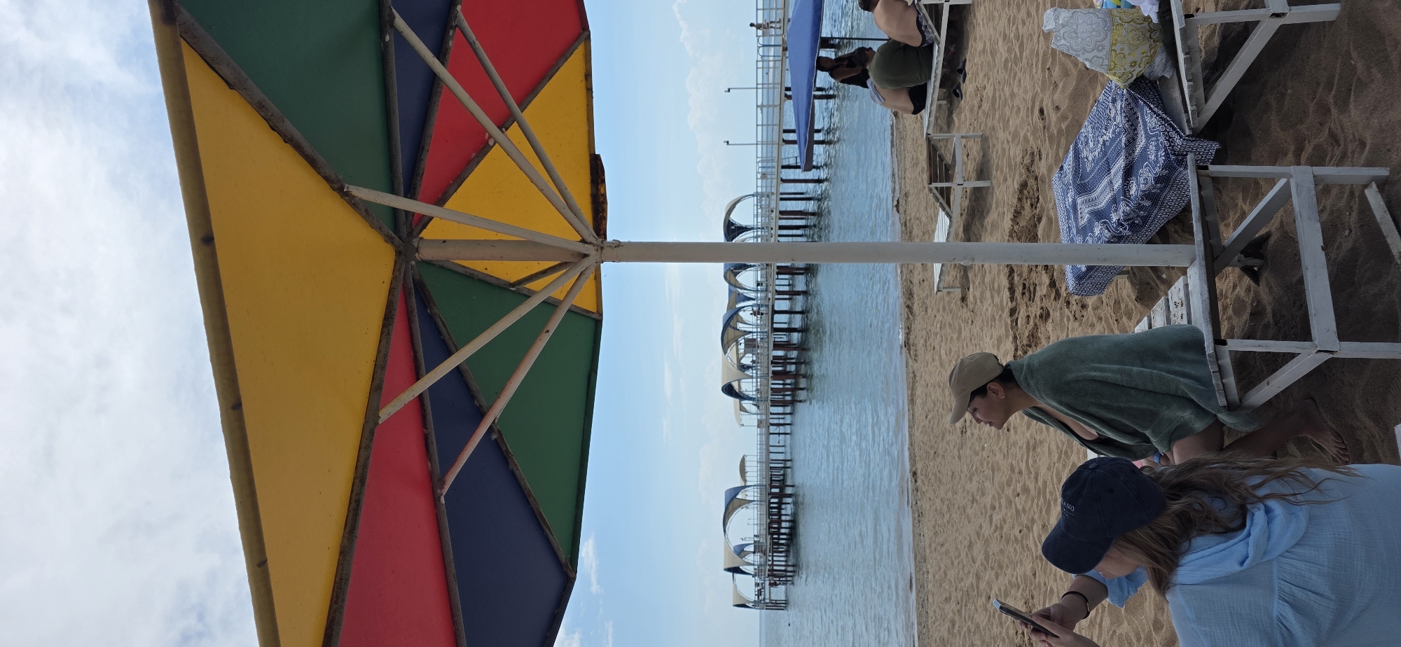 This is a photograph taken on a beach in Kara-Oy, Kyrgyzstan. A large, multi-colored beach umbrella with sections of yellow, red, blue, and green dominates the foreground, obscuring a portion of the sky and the view of the ocean. In the middle ground, a sandy beach leads to the calm, blue water of the Issyk-Kul lake. Several piers with covered seating areas extend out over the water, with a few people visible on them. On the beach in the foreground, two women are seated on a white wooden bench. One woman, wearing a light blue top and a dark blue cap, is looking down at a mobile phone she is holding. The other woman, wearing a beige cap and a dark green towel wrapped around her shoulders, is looking up towards the sky. There are lounge chairs and towels on the sand, and a second pier is visible further down the beach. The sky is partly cloudy.