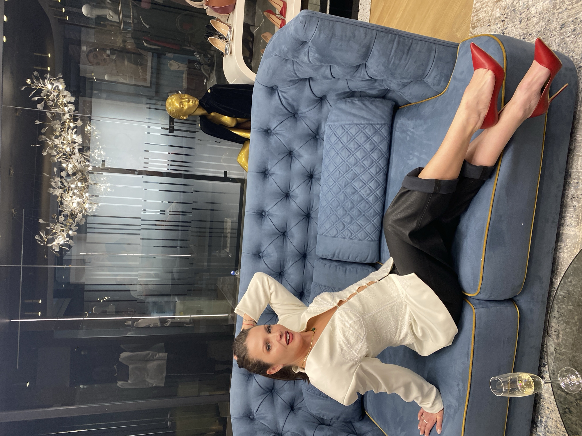 The scene depicts a woman reclining on a blue tufted sofa in what appears to be a high-end retail environment. She is wearing a white top, black pants, and red high-heeled shoes. One arm is raised behind her head. Near the sofa, there's a glass of champagne resting on a table. Behind the sofa is a glass wall or window displaying mannequins and retail displays, suggestive of a store or showroom. The location appears to be Yekaterinburg, Russia.