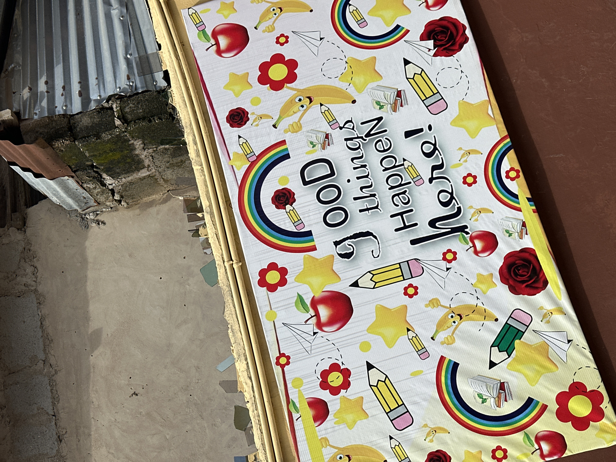 This image presents a vibrant, colorful banner or mat, likely serving as a decorative or instructional piece, set against a rugged, utilitarian background in what appears to be an outdoor or semi-outdoor location in Lagos, Nigeria.
The central subject is a brightly patterned banner featuring various educational and cheerful motifs. The most prominent text reads "Good Things Happen Here!", displayed in large, playful lettering. Surrounding this message are numerous cartoon-style illustrations, including smiling yellow bananas, bright red apples, yellow stars, small red flowers, red roses, paper airplanes, open notebooks, and pencils (both yellow and green). Colorful rainbows also punctuate the design.
The background contrasts sharply with the cheerful banner. To the left, a rough, grey concrete wall made of large, weathered blocks, some covered with green moss, is visible. Above this, two sheets of corrugated metal are attached, one grey and the other a rusty brown, suggesting a functional or unfinished part of a structure. To the right of the banner, a plain, reddish-brown surface, possibly a wall or ground, is partially shown. The immediate area around the banner includes some wooden or bamboo-like supports and fragments of debris on the ground.
The scene appears to be set during the day, indicated by the bright, even lighting, though no strong shadows are cast. There is no specific activity taking place, but the banner's positive message suggests an intention to create an inspiring or welcoming space, possibly for children, within its practical surroundings.