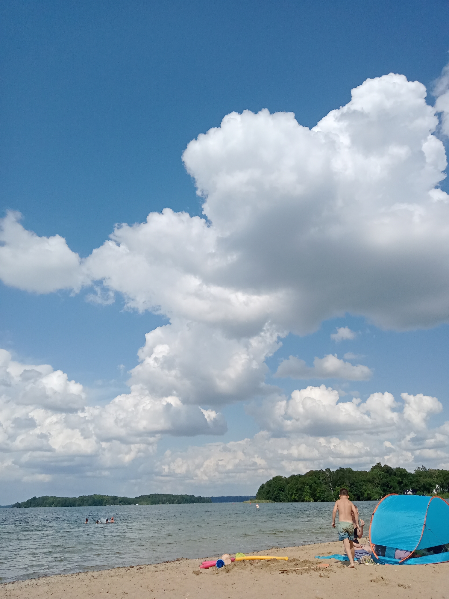 This image depicts a sunny day at a beach, likely located on the shores of the Ziegelsee in Schwerin, Germany. A young boy in swimming trunks is walking away from a bright blue beach tent, heading towards the water. He is seen from behind, with his body facing right and his head turned slightly to the left. To his right, partially obscured by the tent, is another person lying down. Scattered on the sand near the tent are beach toys, including a yellow pool noodle and a pink shovel and bucket. In the water, several people are visible swimming and playing. The lake stretches out to a tree-lined shore in the distance, under a partly cloudy blue sky. The overall atmosphere is one of leisure and outdoor recreation.