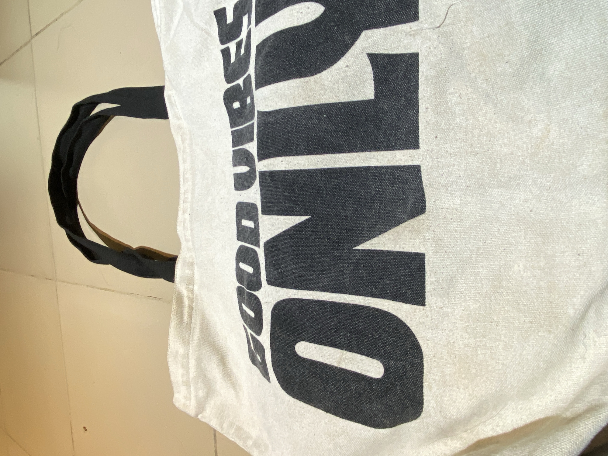 A close-up, slightly angled shot shows a tote bag lying on a light-colored tiled floor. The bag is primarily off-white, made of a canvas-like material, and printed with bold, black lettering that reads "GOOD VIBES ONLY". Black handles are attached to the bag's top. The lettering is prominently displayed, with the "ONLY" section being particularly large and dominating the frame. The tiled floor is visible in the background, providing a sense of the immediate surroundings. No people are visible in the image. The location of the image cannot be determined.