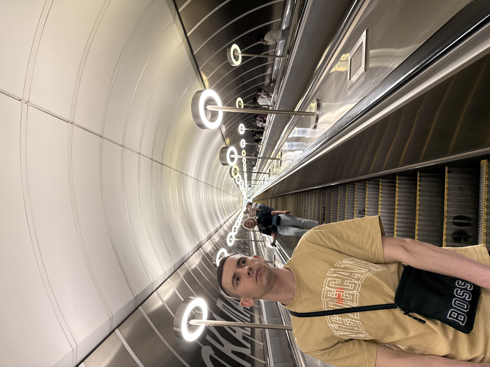 This image captures a long, ascending escalator tunnel within a modern subway station, identified as part of the Moscow Metro. In the foreground, a young adult male stands on the left escalator, slightly turned to his right, looking upwards with a neutral expression. He has short dark hair and wears a mustard-yellow short-sleeved t-shirt with white block lettering, and a black cross-body bag with "BOSS" in white text slung over his left shoulder.
The tunnel features two parallel escalators with black steps, yellow demarcation lines, and dark handrails. The balustrades and lower side walls of the tunnel are highly polished, reflective metallic surfaces. A series of distinctive circular LED light fixtures are mounted on tall, slender poles along the central divider between the escalators, casting bright, cool white light and creating reflections on the metallic surfaces. The upper section of the tunnel wall on the left features large white letters, partially visible as "СКА". The ceiling is a curved, vaulted structure composed of light-colored, segmented panels with subtle linear grooves, extending far into the distance. Several other passengers are visible ascending the escalators further up the tunnel, including a woman in a dark shirt and light jeans on the right escalator. All individuals are standing on the moving escalators.