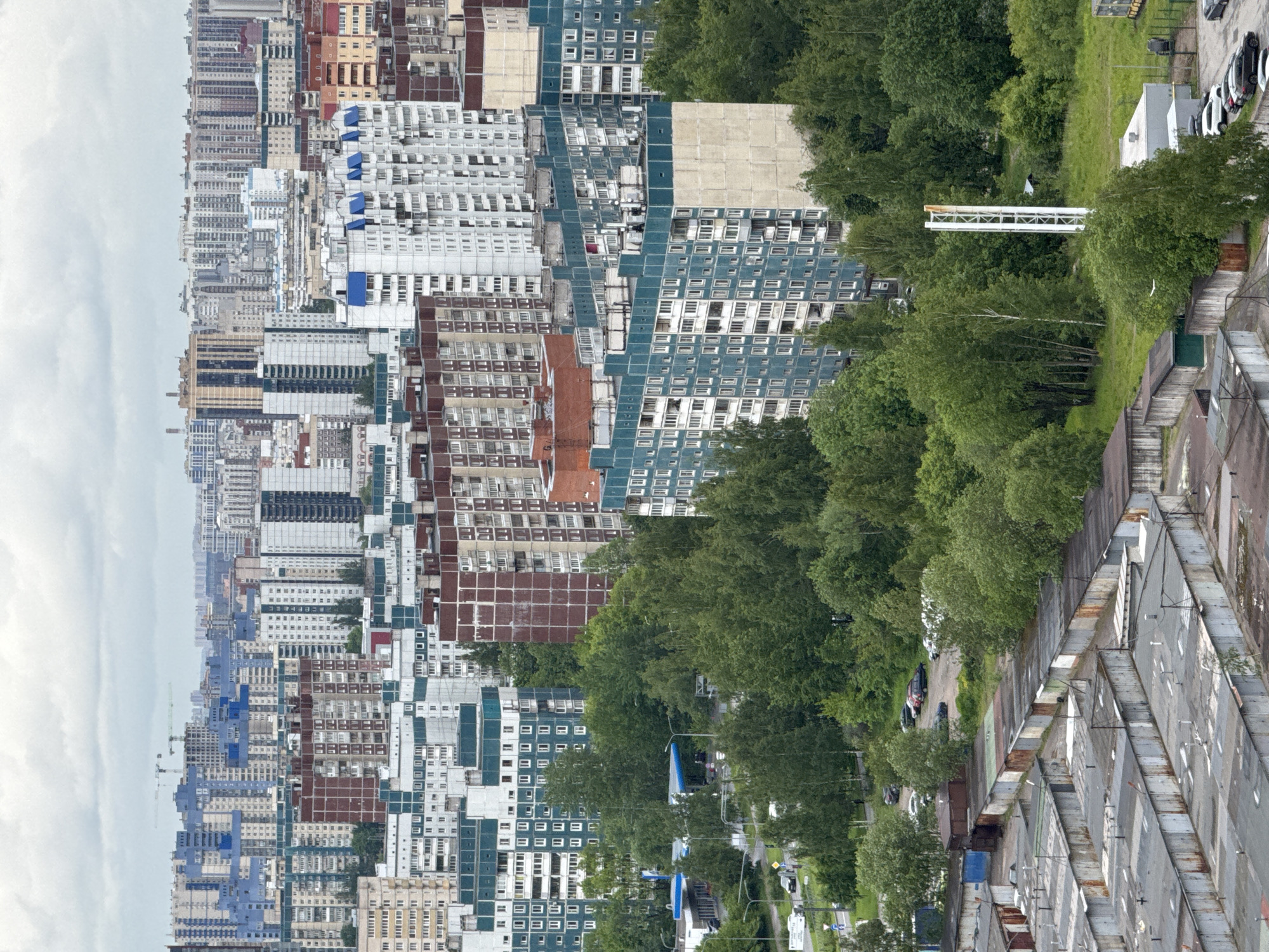 The image shows an aerial view of a cityscape. The city includes rows of high-rise buildings. They are mostly gray, white, and brown. Interspersed with the buildings are patches of green trees and vegetation. A road runs through the trees. Garages or warehouse-like buildings are on the edge of the scene. The location is most likely in Saint Petersburg, Russia, based on the architectural styles and urban planning visible in the photograph.