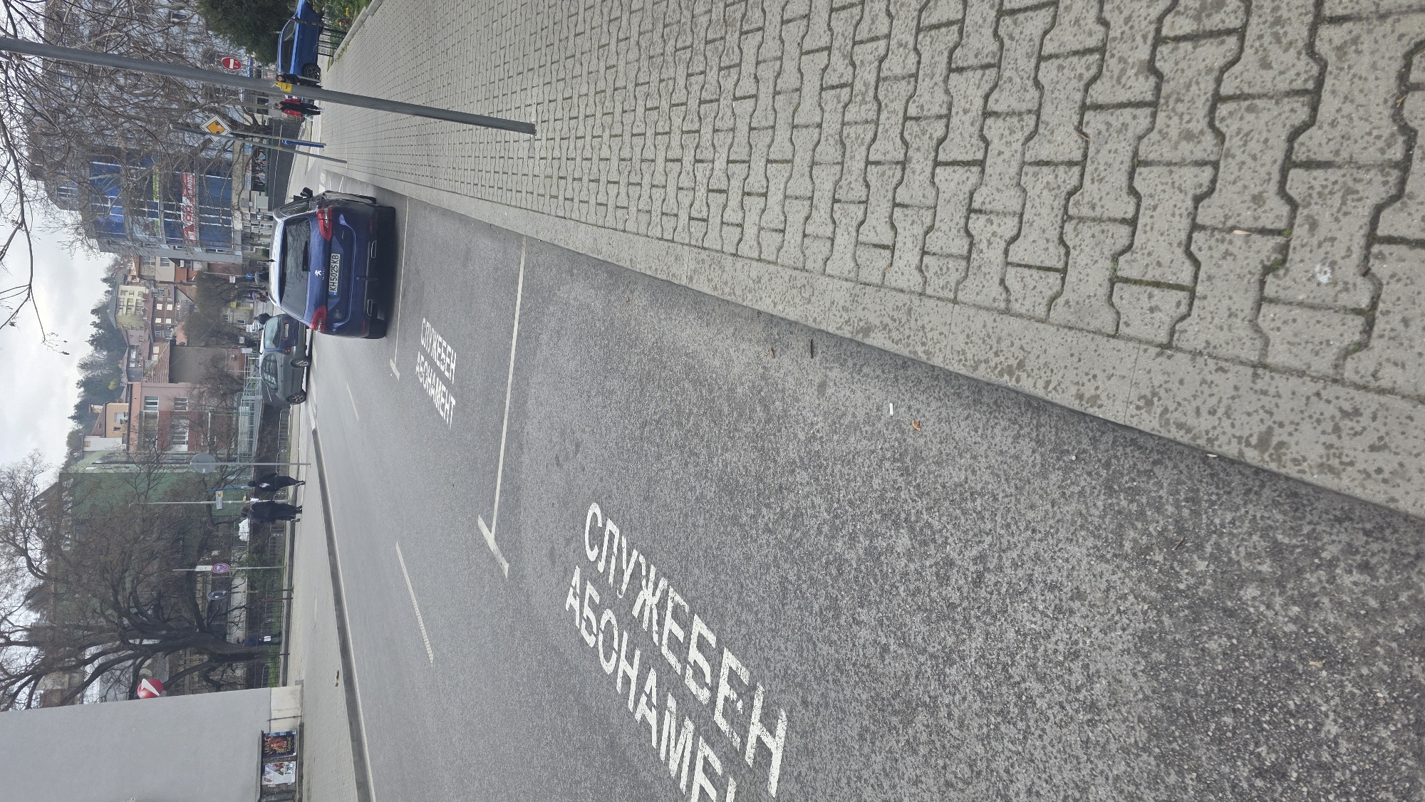 The image captures a street scene in Blagoevgrad, Bulgaria, featuring a parking lane marked with the Cyrillic inscription "СЛУЖЕБЕН АБОНАМЕНТ," which translates to "Official Subscriber." Two parked cars are visible within the designated parking spaces, a dark blue Peugeot 308 SW, license plate KH5025KB, and a gray car in front of it. A pavement composed of interlocking concrete pavers runs alongside the parking lane, leading towards the sidewalk. Several pedestrians are walking on the sidewalk in the distance. The background reveals a cityscape with buildings of varying heights and colors. A tall building with blue and white exterior dominates the scene. Bare trees line the street, indicating the season. A streetlight pole is located on the side of the lane.