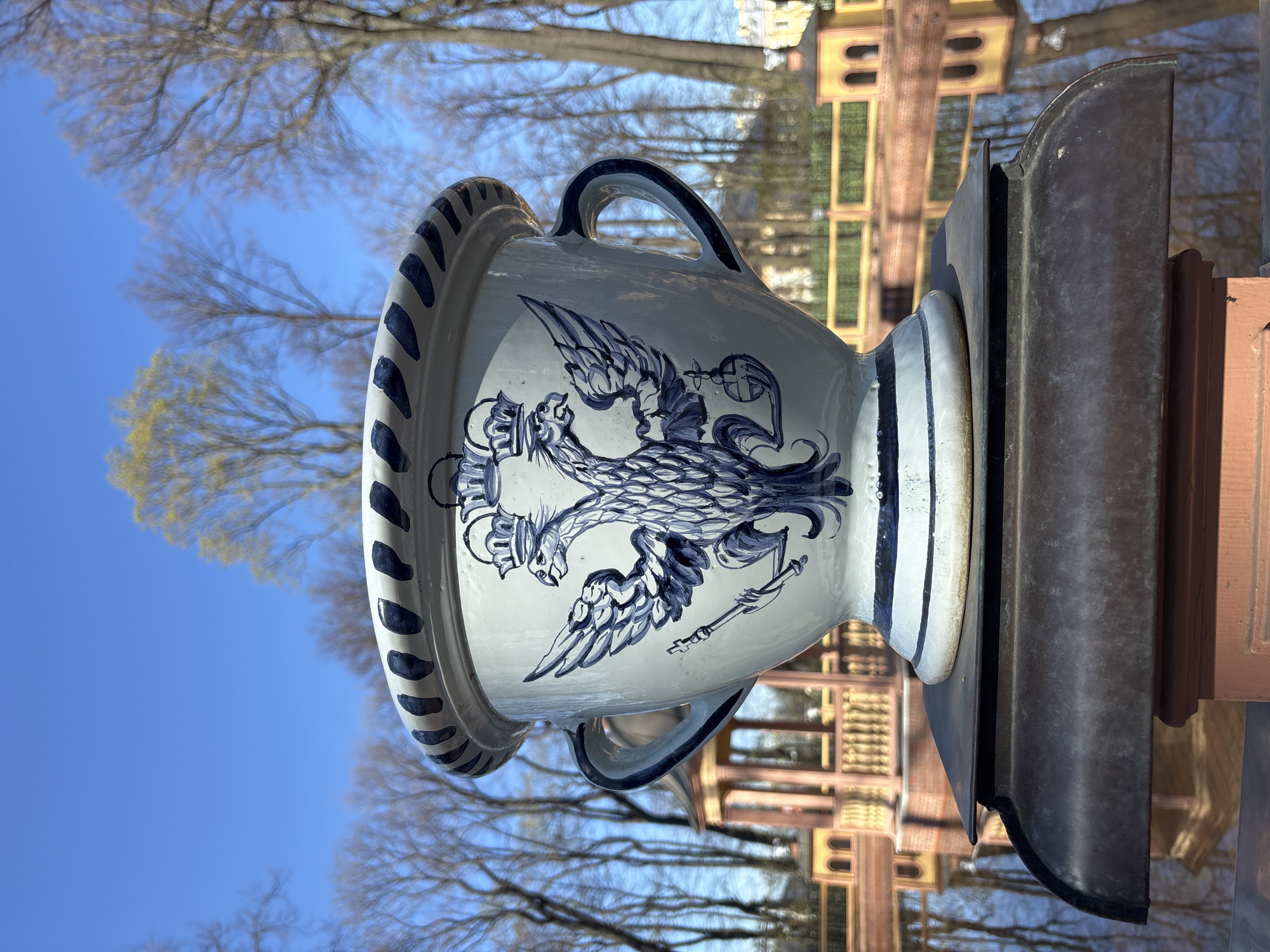 The image shows a large, decorative urn in what appears to be a park in Saint Petersburg, Russia. The urn is predominantly white with blue detailing, featuring a depiction of a double-headed eagle, possibly the Russian coat of arms. It is mounted on a dark, metallic or stone pedestal. The background consists of trees without leaves, suggesting a time either early spring or late fall and a building facade, likely a palace. The sky is a clear blue. The photo is taken in a way that the urn is the focal point. The architecture of the visible structure in the background suggests it could be Peterhof or another similar imperial estate near Saint Petersburg.