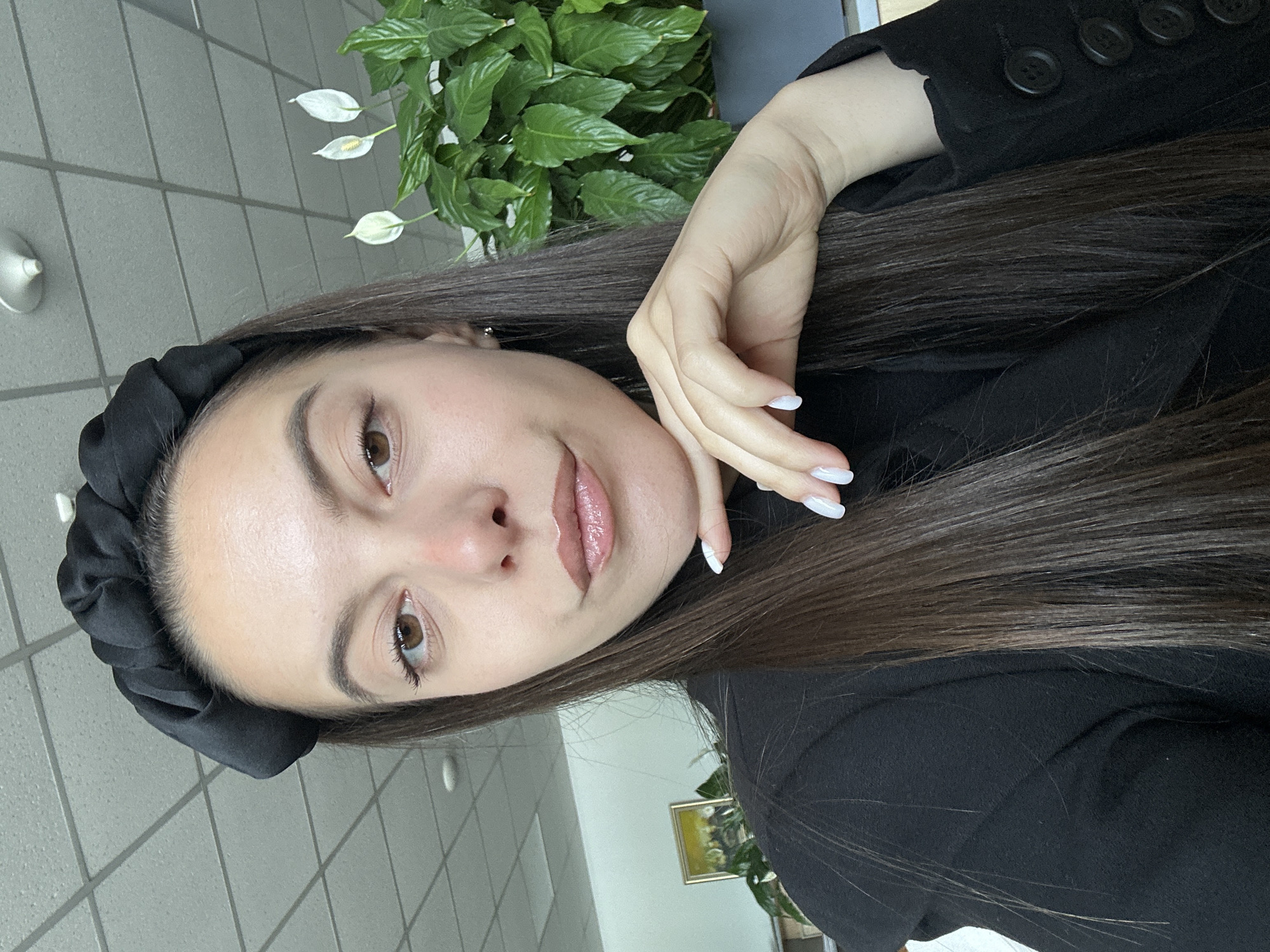 A woman is posing for a selfie in what appears to be an office setting. She has long brown hair and a black head band. She is wearing a black blazer with buttons. Her left hand is near her face, showing light-colored painted nails. A peace lily plant is in the background, along with a tiled wall. The location is possibly in Sofia, Bulgaria.