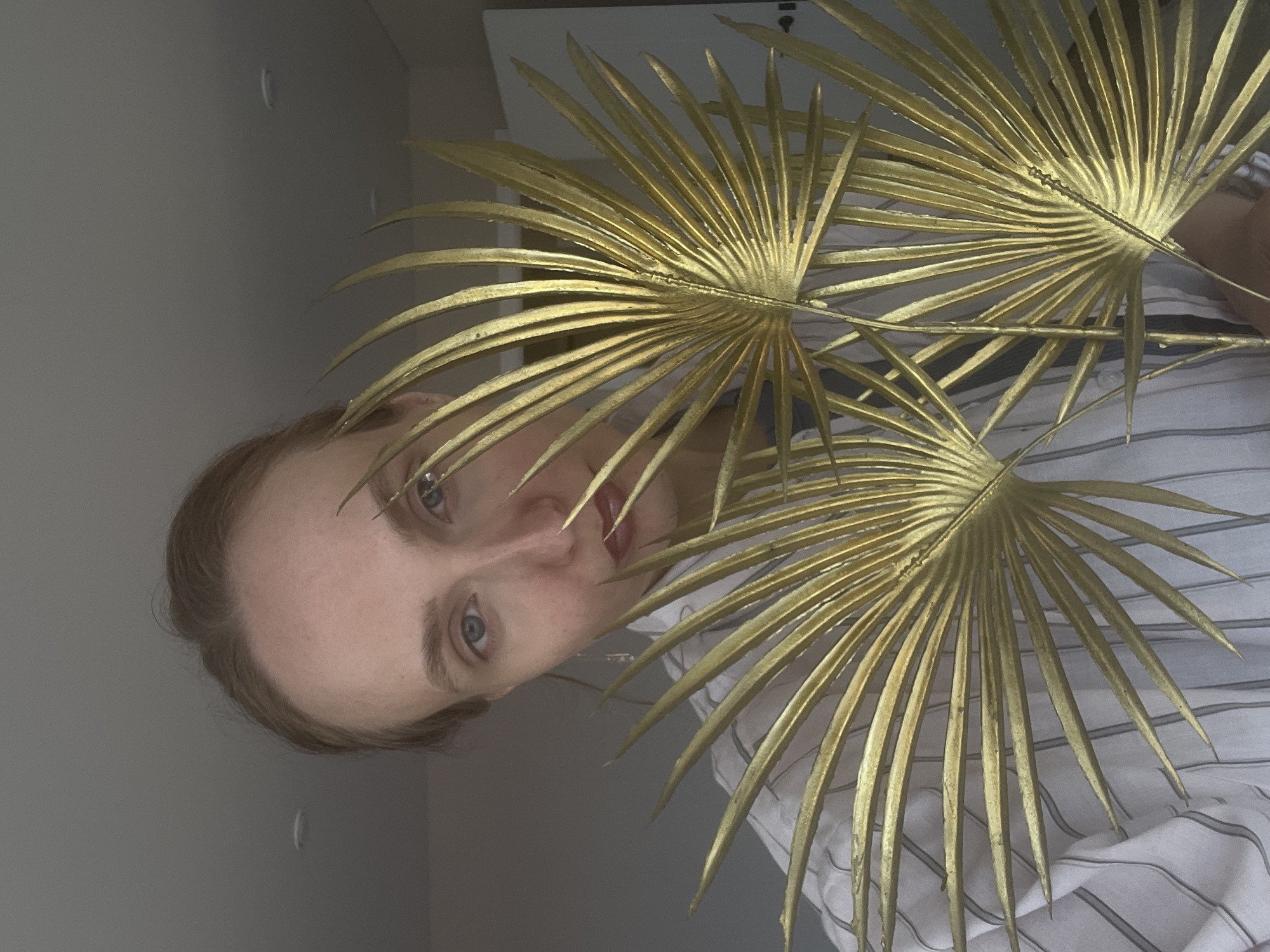 A person with fair skin and light brown hair is posing behind three gold-painted palm leaves. The person is wearing a white and grey striped shirt. The background consists of a grey wall on the left and a white door in the upper center. The photograph has been rotated counter-clockwise. It is not possible to determine the location in the city of Sepych, Russia from the image.