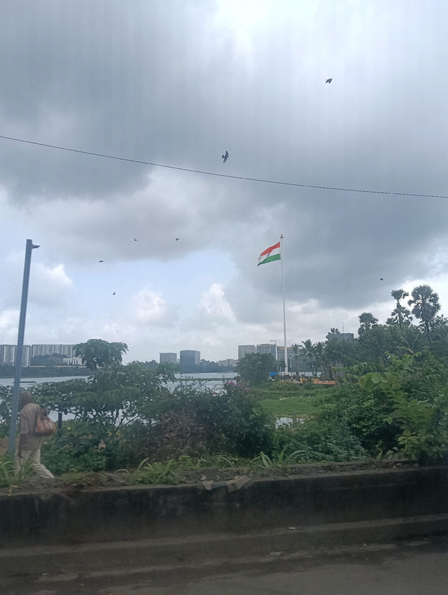 This image captures a view of a cityscape in Mumbai, India, under a cloudy sky. In the foreground, a concrete barrier runs horizontally across the lower portion of the frame. To the left, a person walks from left to right, carrying a bag over their shoulder. Behind them, a tall, gray pole with a light fixture stands vertically.
Further into the scene, a body of water is visible, bordered by lush green trees and vegetation. Across the water, a prominent Indian flag flies high on a tall flagpole, its colors of saffron, white, and green clearly distinguishable. In the background, several modern buildings and high-rise apartments form the city skyline. Several birds are seen flying in the sky. The overall impression is one of an urban environment blended with natural elements, under an overcast sky.