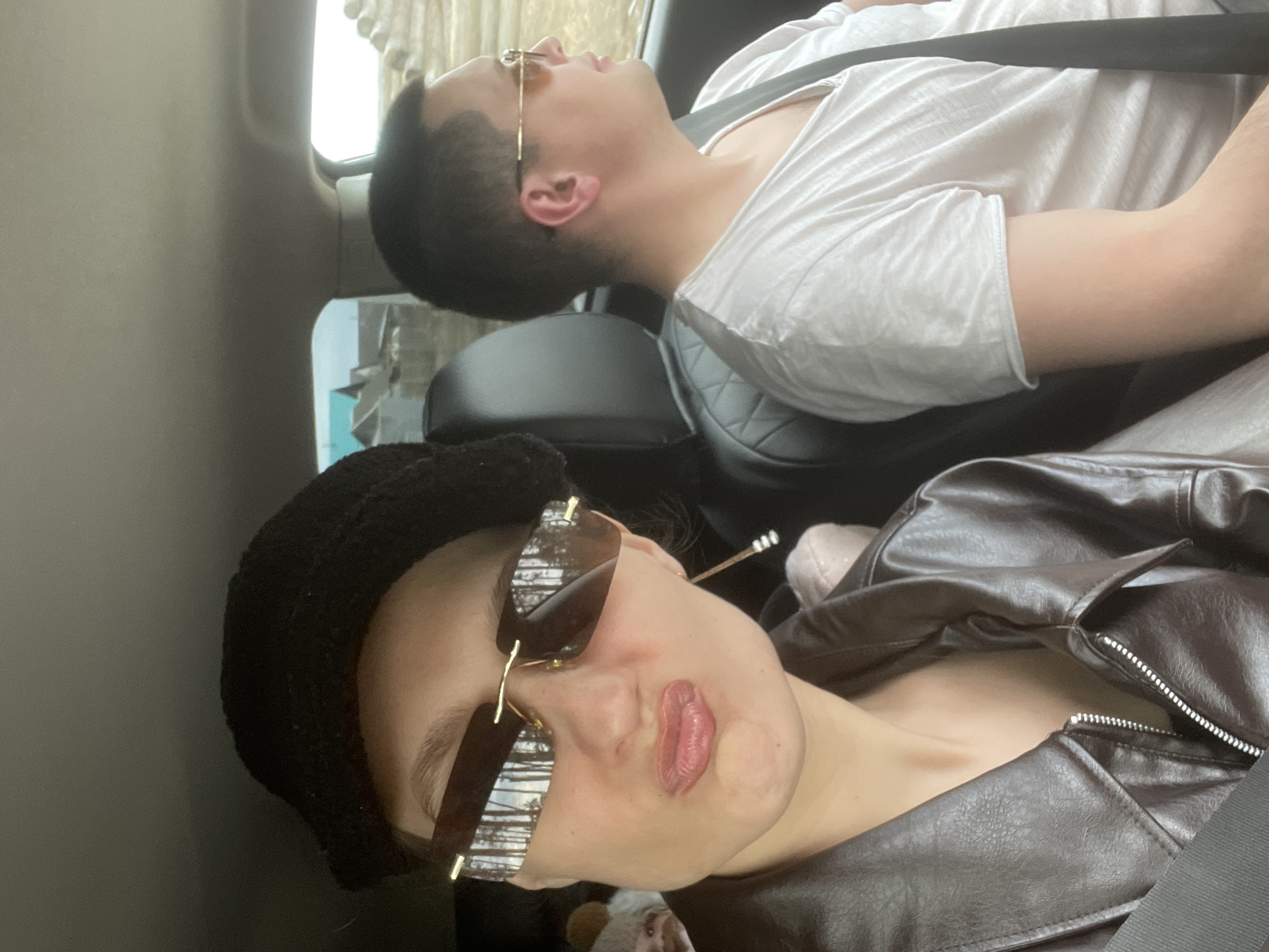 This image, taken inside a car in Подшивалово, Russia, features two individuals during daytime. The perspective is tilted, creating a dynamic view of the interior.
In the foreground, a person is seen wearing a black knit hat, large sunglasses with reflective lenses, and a brown leather jacket. Their lips appear to have lip gloss or lipstick. They are facing towards the camera with a neutral expression, largely obscured by the sunglasses.
Further back in the vehicle, a second individual is visible, resting or possibly asleep. They are wearing a white t-shirt and gold-rimmed glasses, secured by a seatbelt. Both subjects are seated in car seats.
Bright daylight illuminates the car interior through the windows. Reflections in the foreground person's sunglasses offer blurry glimpses of an outdoor scene, possibly trees or distant structures, suggesting travel through a rural or suburban environment. The overall activity depicted is car travel. No visible text is present in the image.