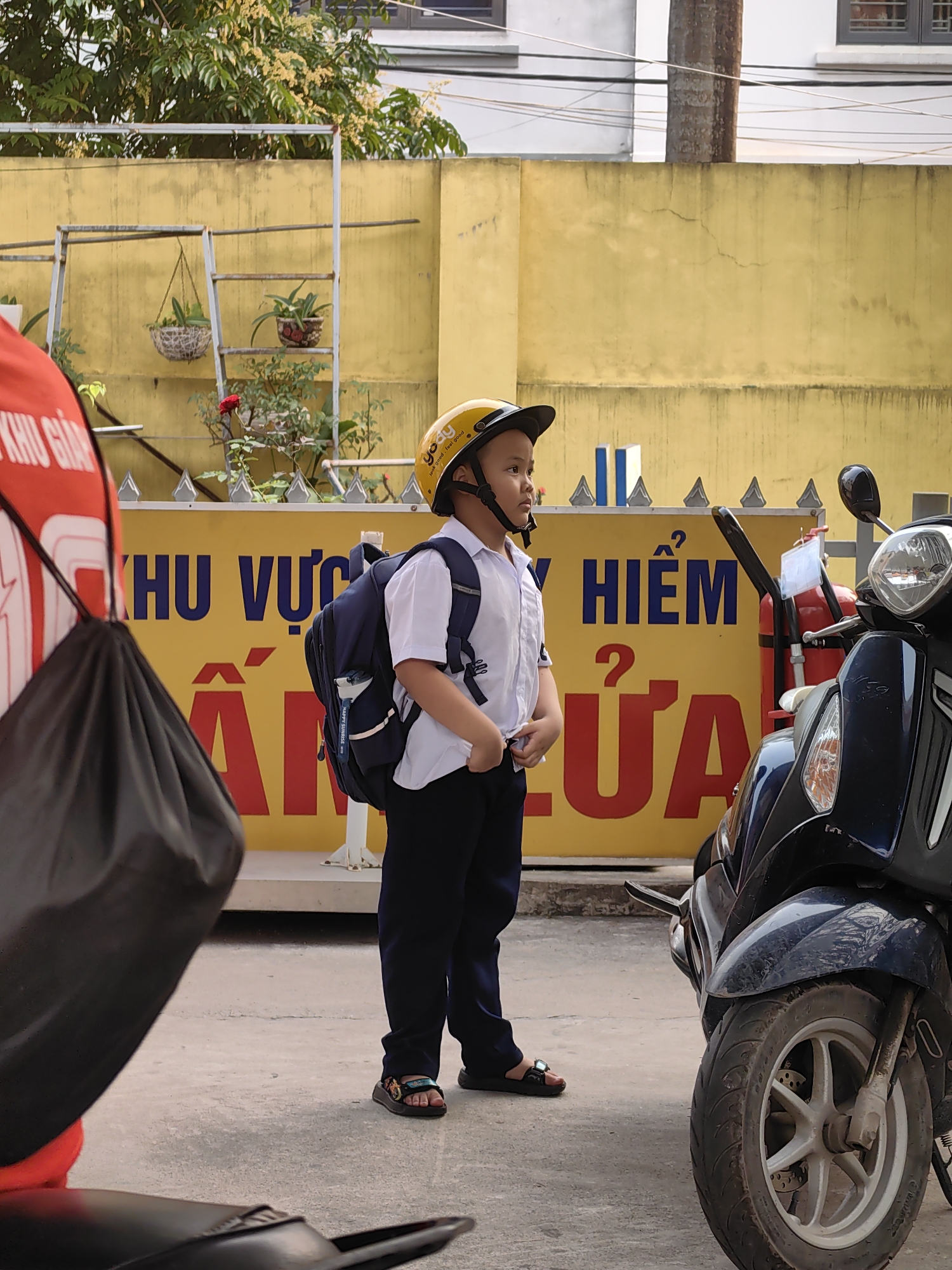 A young boy wearing a white short-sleeved shirt, navy pants, sandals, and a yellow helmet with a black visor is standing in front of a yellow wall. He has a blue backpack on. The sign on the yellow wall behind him is in Vietnamese, with large blue and red text. To the boy's right is a dark blue scooter. Partially visible to the left is a person wearing a red shirt and carrying a black bag. A metal ladder with potted plants is visible on top of the yellow wall. The setting is a street in Ha Lam Ward, Vietnam.