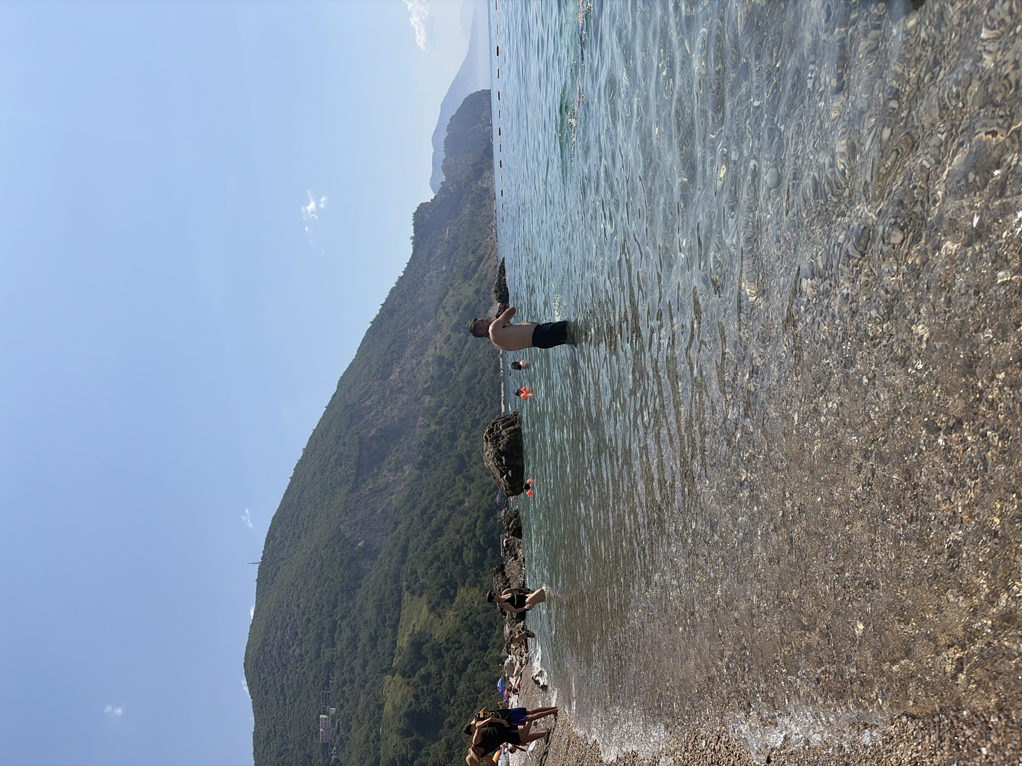 This is a wide-angle shot of a pebble beach in Prijevor, Montenegro. The foreground is dominated by the clear, shallow water lapping onto the shore, revealing the colorful pebbles beneath the surface. In the middle ground, several people are visible in the water. A man with dark hair and dark shorts stands chest-deep in the sea, facing away from the camera. To his left, a woman in a black swimsuit is also in the water, facing the shore. Further back, several other individuals are present in the water, some wearing orange life vests. The shoreline to the left is lined with a few beach umbrellas and groups of people. The background features a steep, tree-covered mountain range that slopes down towards the sea. The sky is a clear, bright blue.