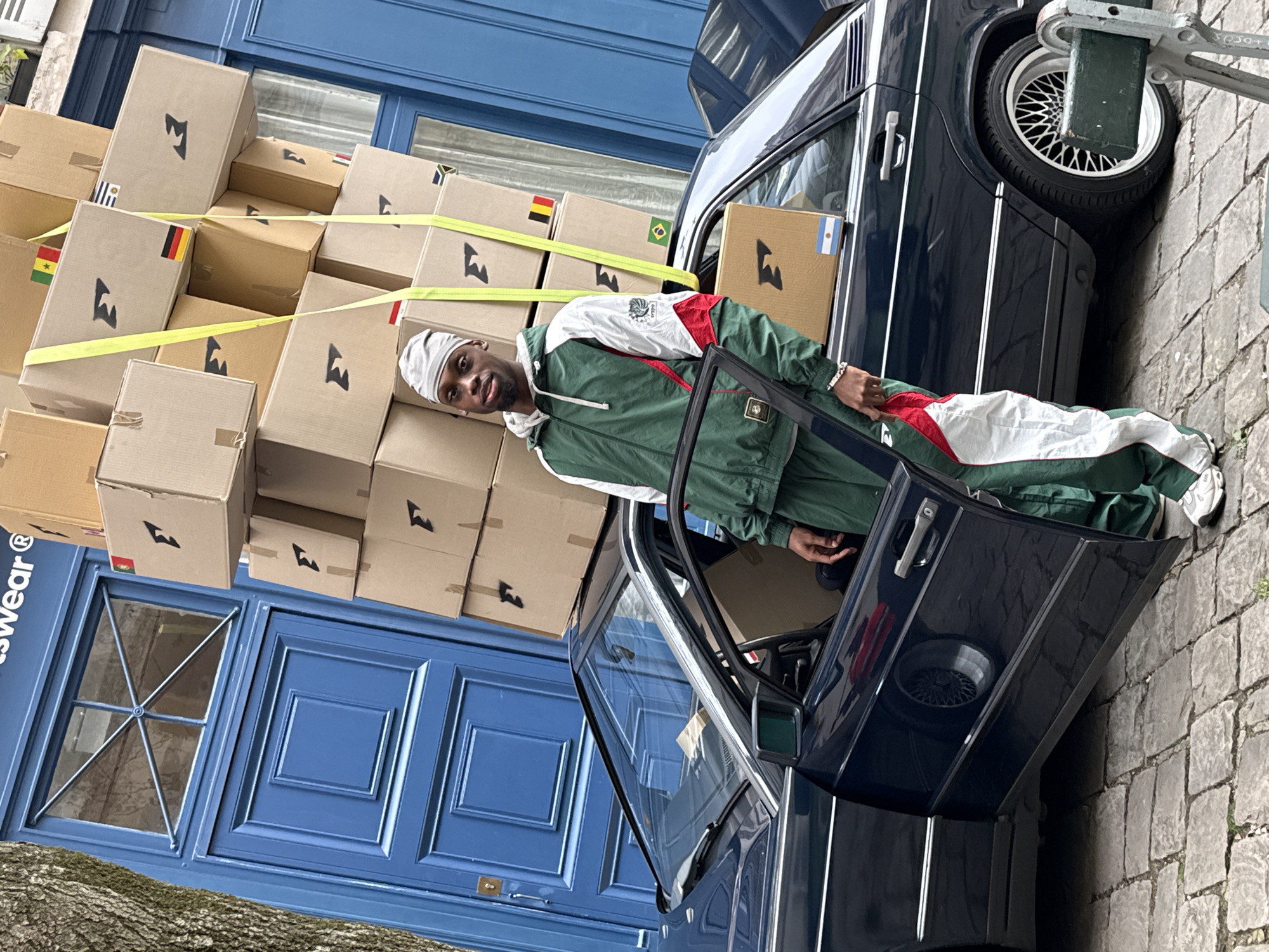 A man wearing a green and white tracksuit is standing next to a dark blue car. The car's door is open and boxes are stacked on top of the car, secured with yellow straps. The boxes are all marked with a logo. Some boxes have flags on them (Ghana, Germany, Brazil, Argentina). In the background is a blue building with windows. The image was taken outside a store with the word "swear" on the storefront in Paris, France.