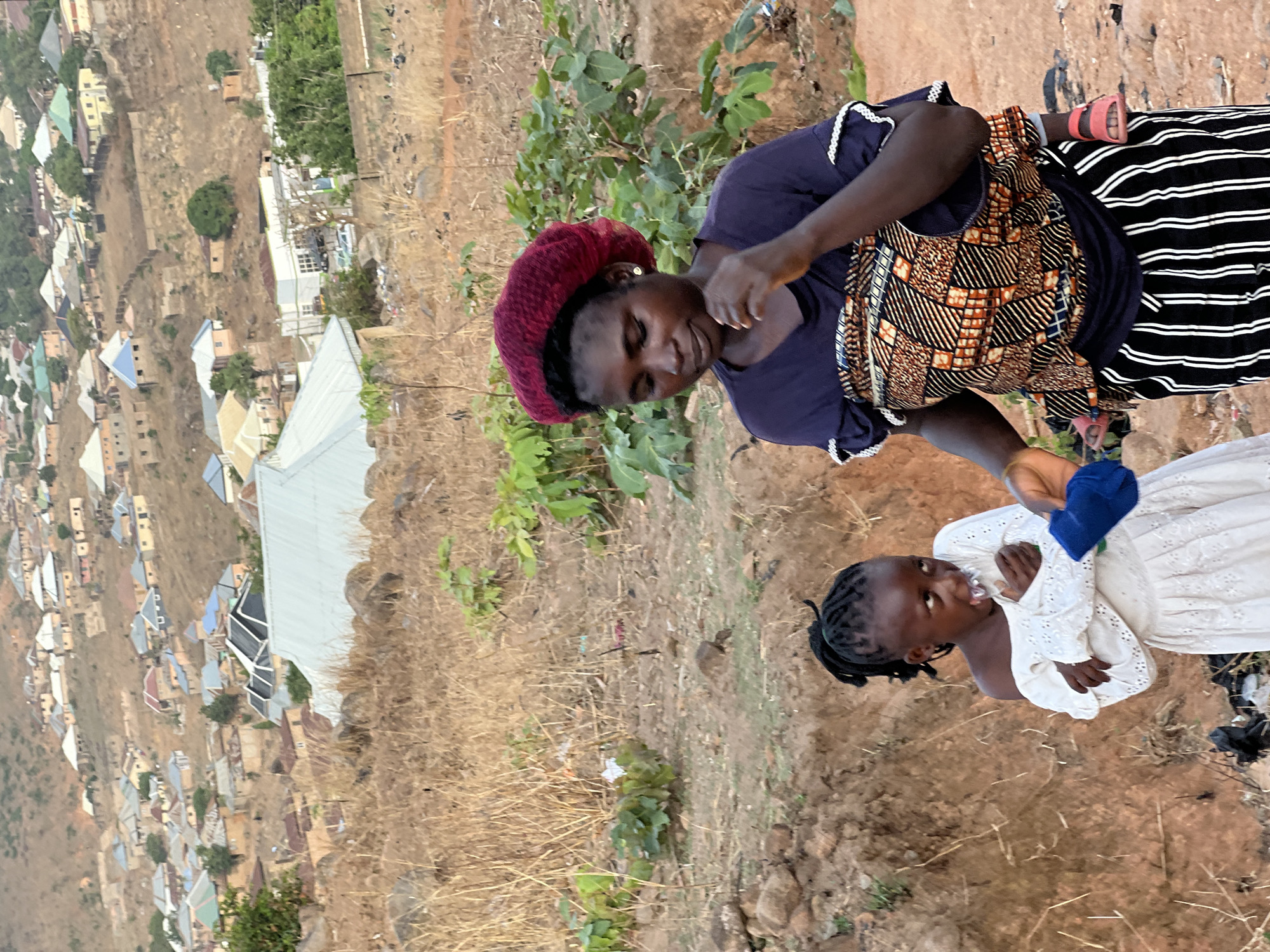 A medium shot depicts a dark-skinned woman in a maroon cap, navy blue shirt with a patterned orange, brown, and black design, and a black and white striped skirt, appearing to interact with a young dark-skinned girl in a white dress and a blue sock. The interaction seems focused, possibly involving clothing or care. They are positioned on uneven, dry, dirt ground with scattered vegetation. The background reveals a landscape view of Jos, Nigeria, characterized by densely packed buildings with diverse roofing styles, indicative of an urban or suburban setting. The scene is shot from an elevated angle, suggesting a vantage point above the subjects and the cityscape.