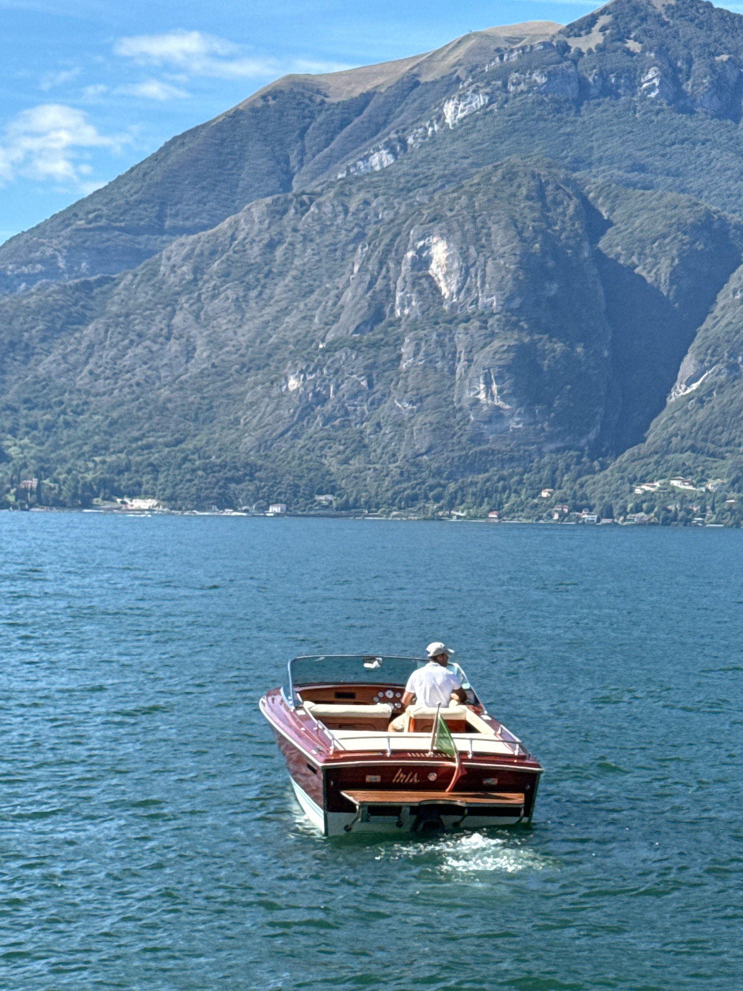 In Varenna, Italy, a classic wooden boat with a rich mahogany hull and white trim navigates the clear, blue waters of a lake. The boat has a single pilot wearing a white shirt and a light-colored baseball cap, seated at the helm. Behind the pilot, there are plush white seats. A small Italian flag is affixed to the stern of the boat. The boat leaves a foamy wake behind it. In the background, the rugged, tree-covered mountains of the Italian Alps rise dramatically, their slopes meeting the bright blue sky dotted with wispy clouds. The shoreline in the distance features scattered buildings and dense green foliage.