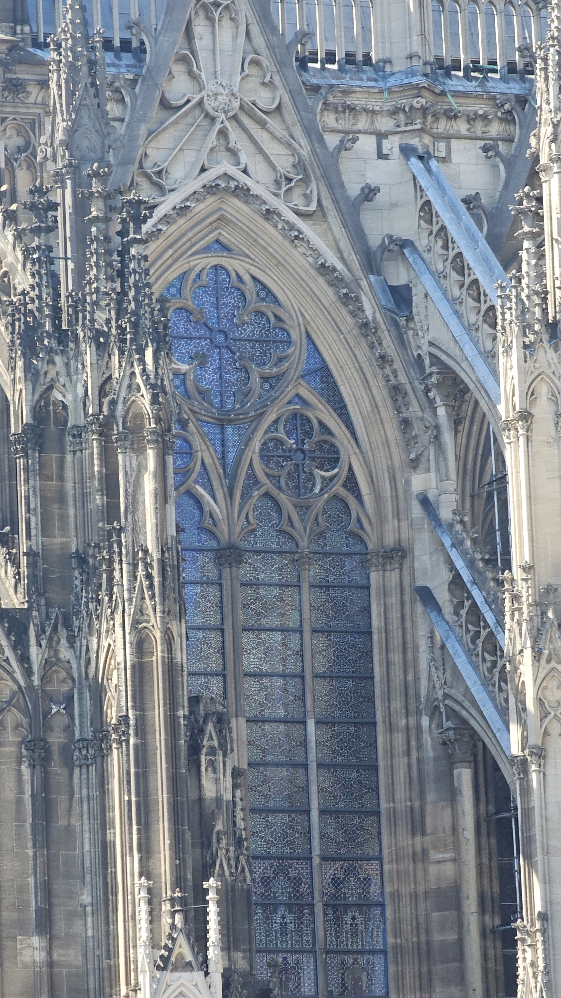 The image is a close-up, angled shot of the exterior of Cologne Cathedral in Cologne, Germany, focusing on a large, ornate stained-glass window. The window is pointed-arch shaped and features intricate tracery with blue and patterned glass. The surrounding architecture is Gothic, with stone buttresses, pinnacles, and decorative carvings, all rendered in shades of gray and beige stone. Sunlight illuminates the facade, casting shadows and highlighting the architectural details. No people are visible in the frame.