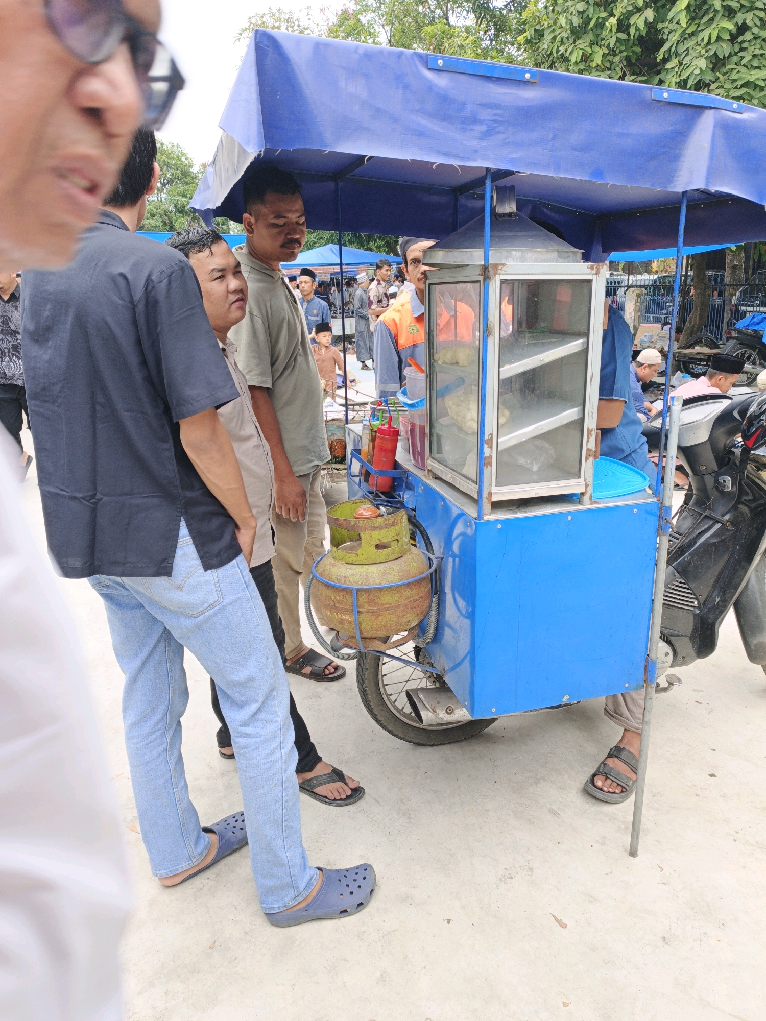 In Manunggal, Indonesia, a street food cart is set up under a blue canopy. The cart, painted bright blue, is equipped with a glass display case showing food items, a stove, and two green propane tanks. A motorcycle is parked to the right of the cart, its front wheel visible. Several men are gathered around the cart, some appearing to be customers and others potentially vendors. One man, wearing a black shirt and light blue jeans, stands with his hands in his pockets. Another man, in a light green shirt, faces forward, looking towards the cart. A third man, wearing an orange and gray vest over a blue shirt, stands behind the cart, seemingly tending to the food. The ground is a paved surface, and trees are visible in the background. The overall scene suggests a casual, outdoor market or food stall environment.