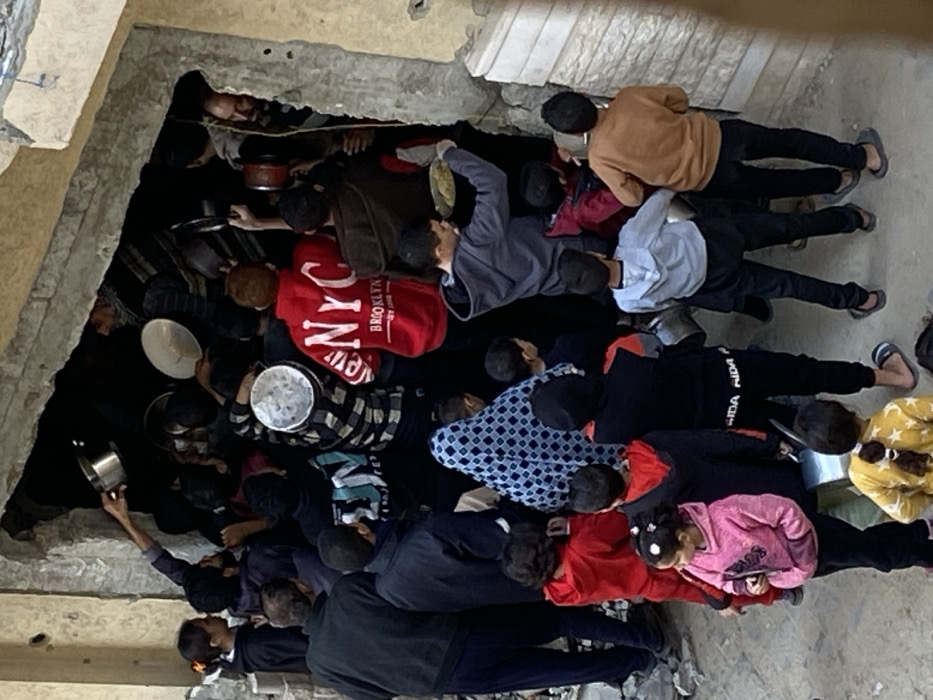 The image displays a dense gathering of approximately 30-40 individuals, including men, women, and children of various ages, concentrated around an irregular, roughly broken opening in a concrete wall, serving as a building entrance, in Khan Yunis, Palestinian Territory. Numerous individuals are holding metal pots, pans, and shallow bowls, some appearing empty, while others contain a light yellow, granular substance, consistent with rice or similar grains. People are actively reaching and pushing towards or through the opening. A young male in a grey hooded sweatshirt is visible holding a yellow plate containing food. Another male figure inside the opening extends an empty silver pot outwards. The immediate ground level in the foreground appears rough, and the adjacent wall section to the right of the opening is light-colored and shows signs of damage. The scene depicts a concentrated distribution or collection of food.