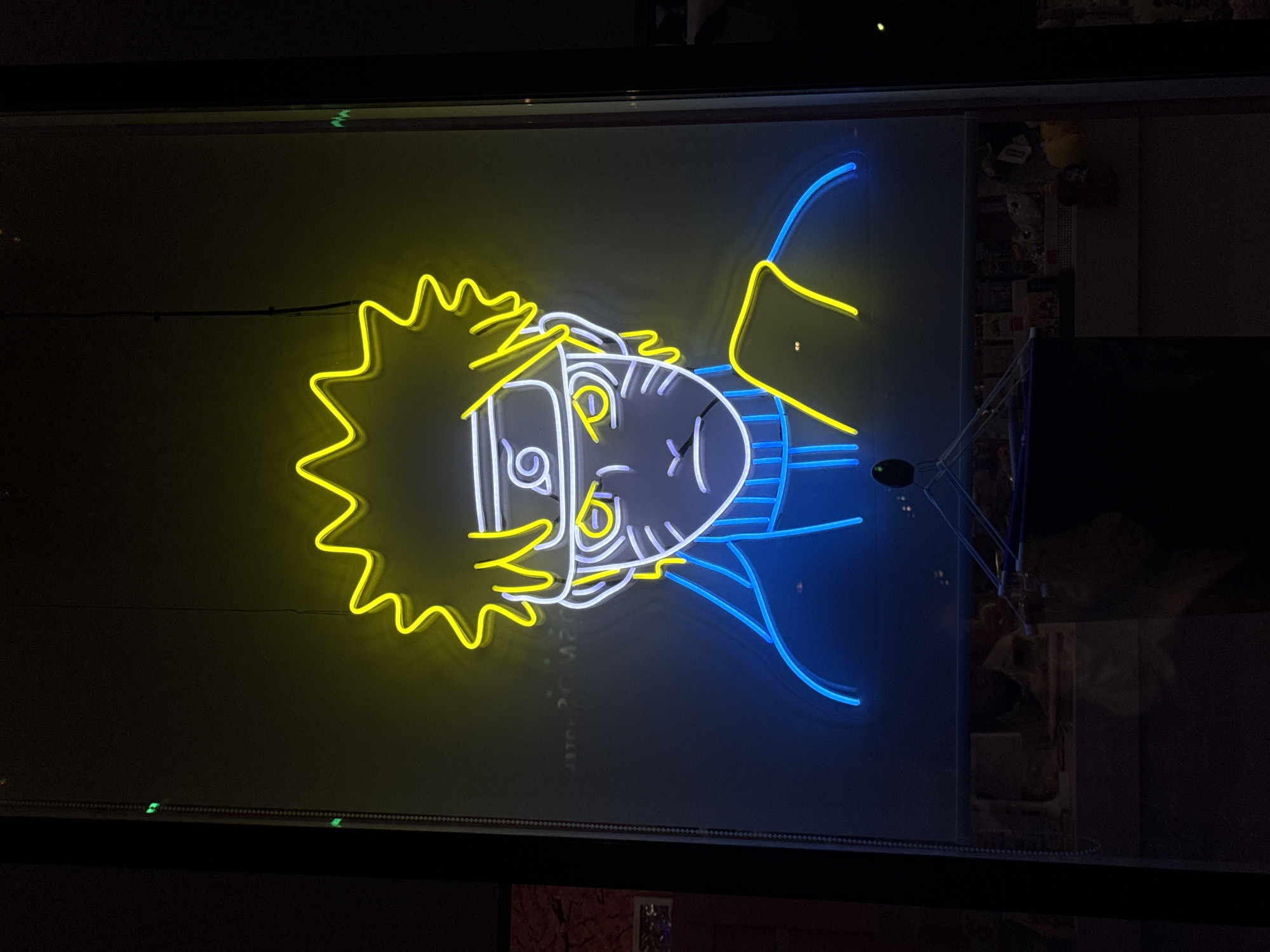 The image depicts a neon sign of Naruto Uzumaki, a character from the anime series *Naruto*. The sign is predominantly yellow and blue, with some white accents. It appears to be displayed behind glass, possibly in a store window or display case. The location is Unknown, Unknown. The rest of the surroundings are dimly lit and indistinct. A stand with a microphone is reflected in the window.