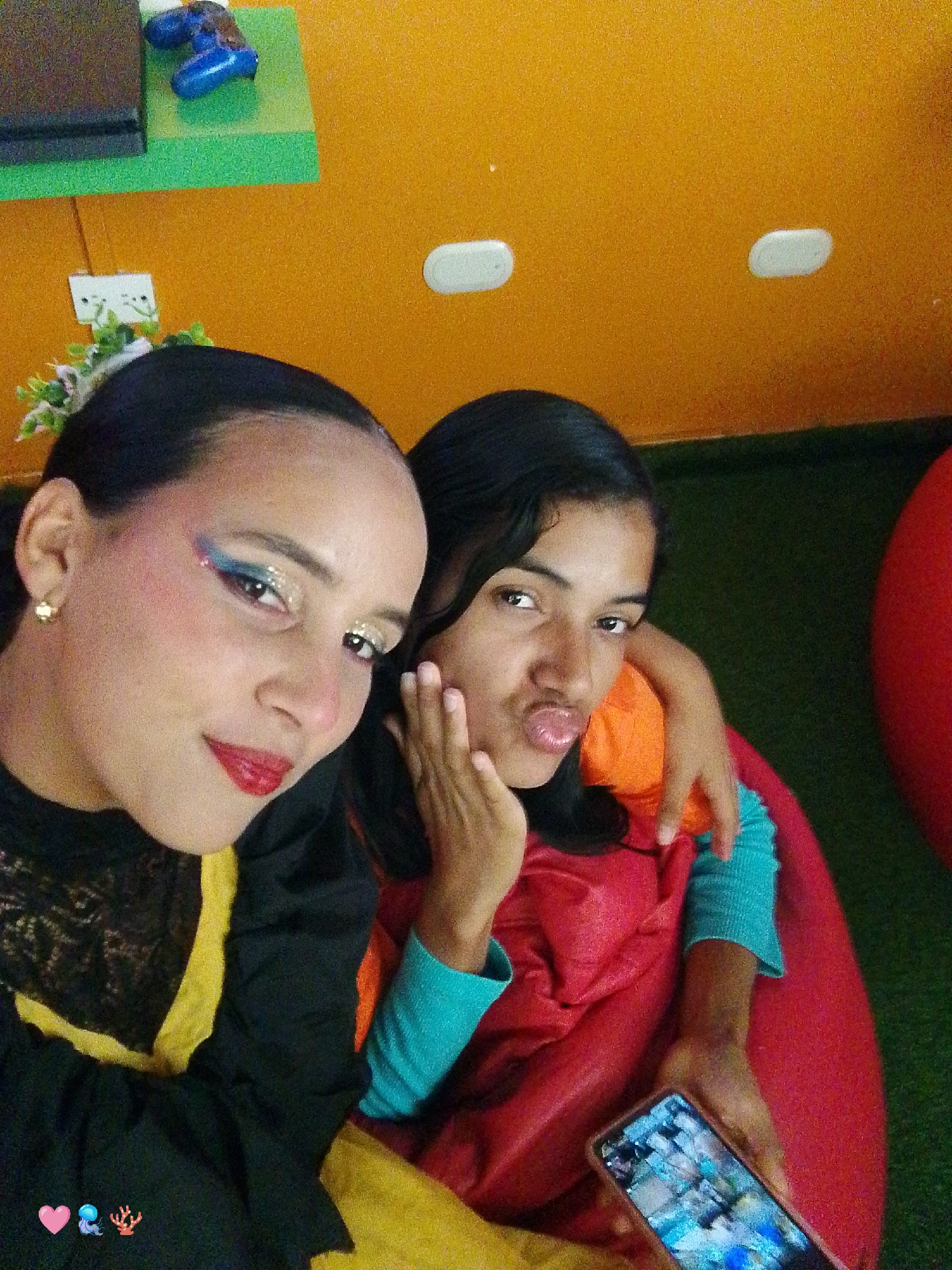 Two young women are in a selfie, indoors. The woman on the left has dark hair, floral decoration in her hair, heavy makeup with blue and silver eyeshadow and red lipstick, and is wearing a black dress with lace and yellow trim. The woman on the right is puckering her lips and sticking out her tongue. She has one hand on her cheek and the other is holding a smartphone. She's wearing a turquoise shirt underneath a red garment. They are both sitting in a red beanbag chair. The background consists of an orange wall with white recessed lights, green astroturf, and what appears to be a shelf with a black box and blue game controller. Based on online search results, this photo may have been taken in a play area or public space in the city of Pedro González, Venezuela.