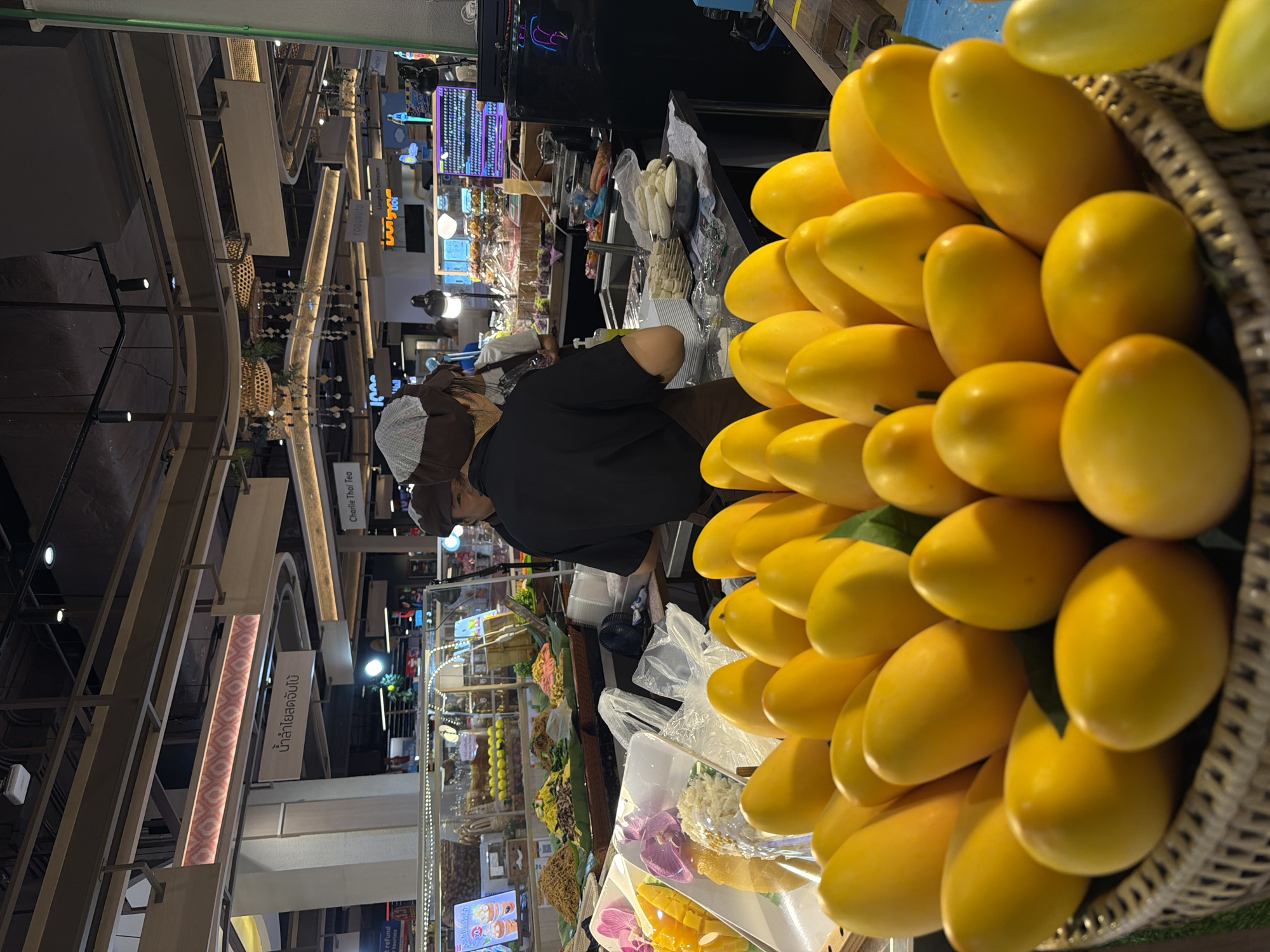 A person is working at a food stall in Chiang Mai City Municipality, Thailand. A large basket of yellow mangoes is displayed prominently on the right side of the frame, taking up a significant portion of the view. The person is wearing a black shirt and a brown cap, facing toward the stall with other food items visible. Behind the stall, there are displays and signs in Thai. The environment looks like an indoor marketplace. Overhead lighting and decorative elements indicate a commercial space. Other customers and food items are visible in the background.