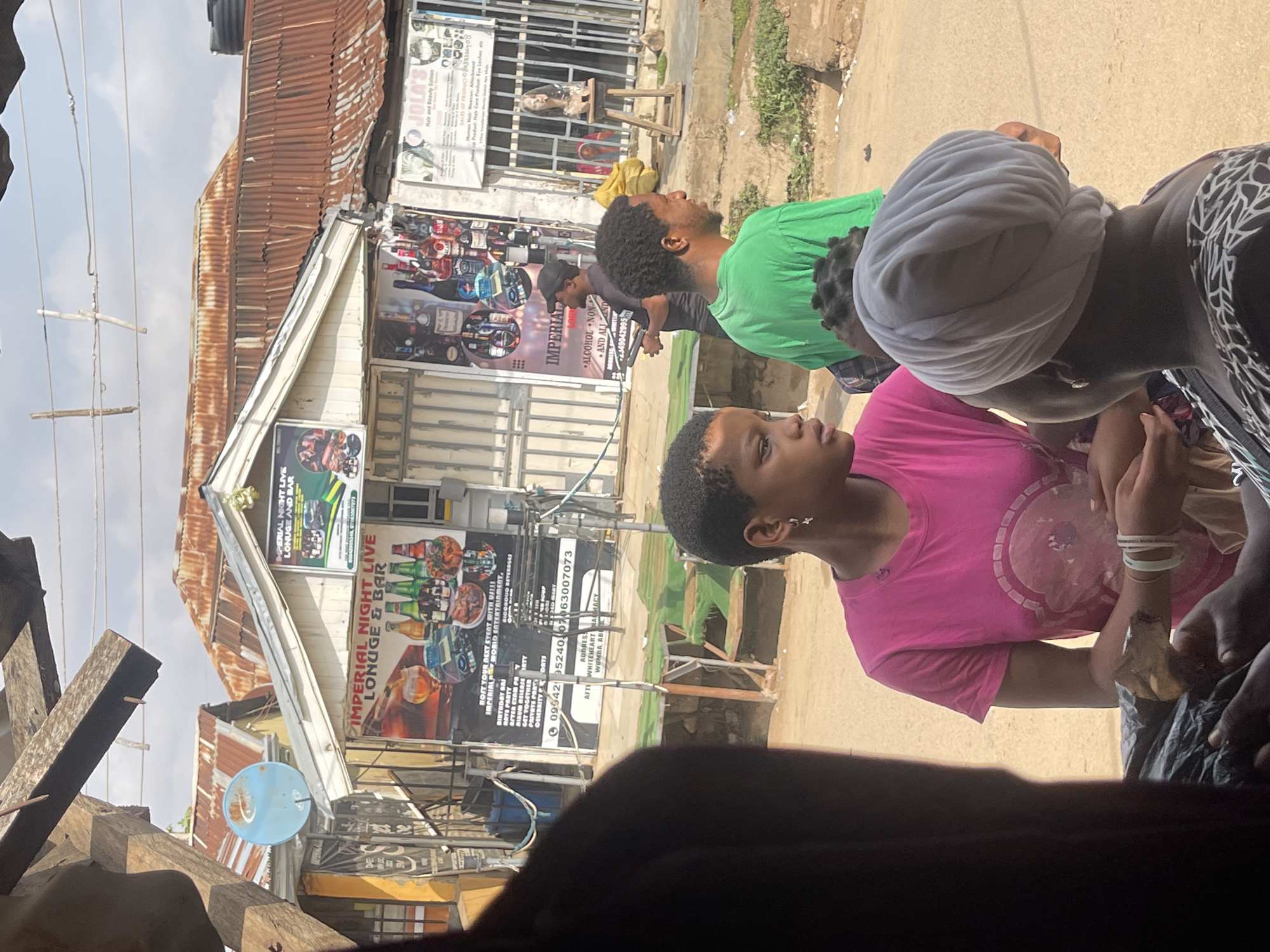 The image shows a street scene in what appears to be Abuja, Nigeria. The view is partially obstructed by a dark foreground object. Three people are visible: one in a pink shirt, another in green, and a third with a white head covering. An "Imperial Night Live Louge & Bar" sign is visible, along with another sign for "Jola's Hair and Beauty Salon." The signs indicate that the establishment offers alcoholic beverages and beauty services. The buildings are made of corrugated metal. A satellite dish is attached to one of the buildings.