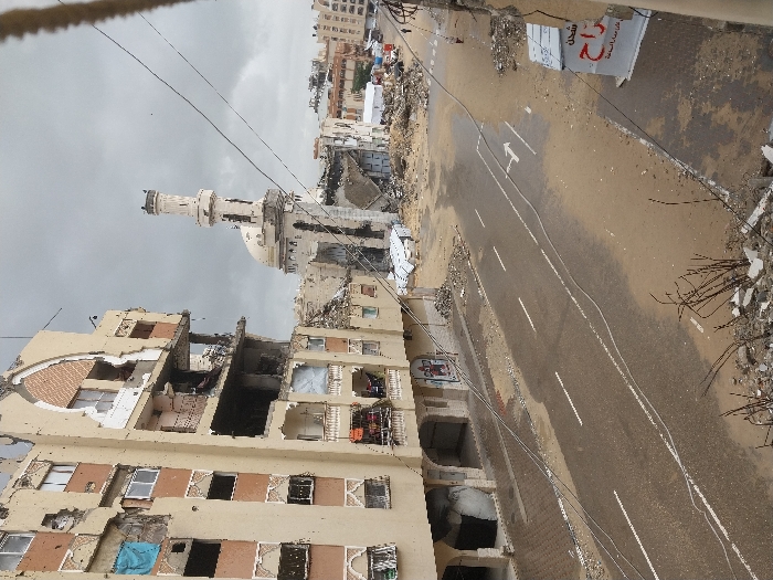 This image depicts a scene of extensive urban devastation in Khan Yunis, Palestinian Territory, under an overcast sky.
In the foreground, debris including broken concrete and bare plant stalks lies on a raised edge, overlooking a two-lane asphalt road. The road surface is partially wet and covered with sand and mud, displaying white dashed and solid lane markings, with a white arrow indicating a turn in one lane. Overhead, multiple utility lines traverse the scene diagonally.
To the immediate left, a multi-story building with a light-colored facade exhibits significant structural damage across multiple floors. Large sections of its exterior walls are missing, revealing exposed interiors, including visible rooms and balconies with ornate railings, some of which are collapsed or damaged. Blue fabric or tarp covers a window opening on one of the upper floors. The ground floor features shopfronts, one displaying a faded mural or graffiti.
Further into the background, prominently positioned in the center-right, is a heavily damaged mosque. It features a visible dome that appears partially collapsed or pockmarked, and a tall, slender minaret that, while standing, shows clear signs of damage. The area immediately surrounding the mosque is a field of rubble and collapsed structures.
To the far right, partially visible, are additional damaged buildings and a temporary white tent-like structure. A white sign with Arabic text, "شحن" (shipping/charging) and "راح" (went/will go, or part of a longer word), is affixed to a damaged structure on the right edge of the frame. One individual is discernible walking in the distance on the right side of the street, and a red vehicle is visible among the debris further down the road.